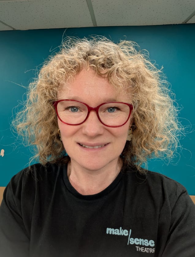 Smiling woman with curly blonde hair and red glasses wearing a black 'make sense THEATRE' t-shirt against a teal wall background.