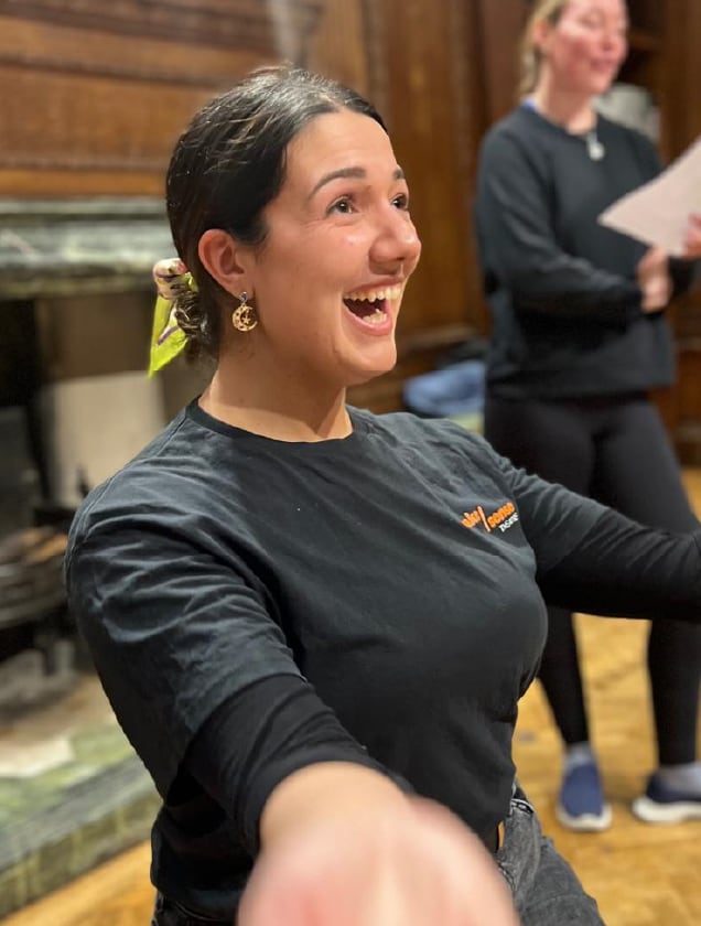 Smiling woman with dark hair tied back, wearing a black shirt with an orange logo, raising her arms and smiling