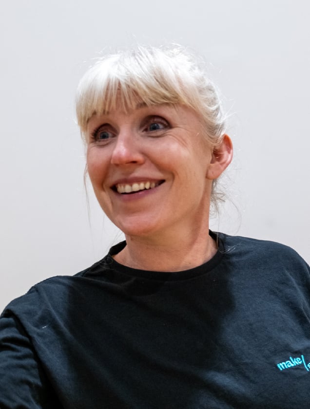 Smiling woman with blonde hair tied back wearing a black shirt against a plain background.