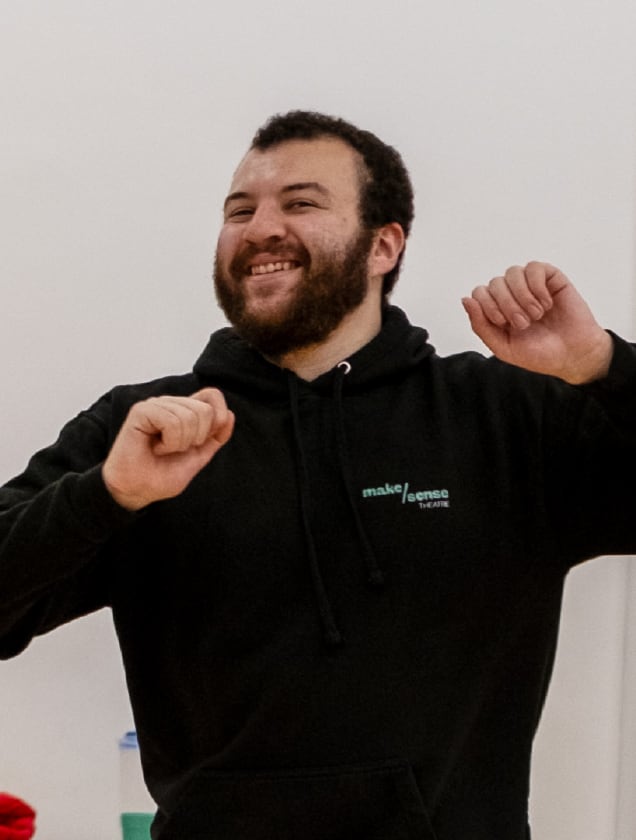 Smiling man with beard wearing a black hoodie raising both hands in a cheerful gesture.