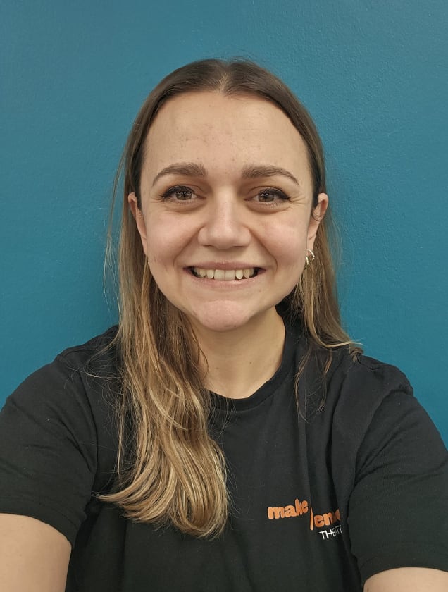 Smiling woman with long blonde hair wearing a black t-shirt in front of a blue wall.