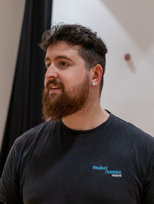 Bearded man with short curly hair wearing a black Make Sense Theatre t-shirt and a stud earring looking to the side.