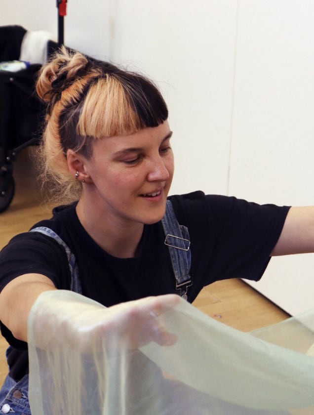 Young woman with half blonde, half black hair tied in a bun, wearing a black shirt and denim overalls, holding a translucent fabric.