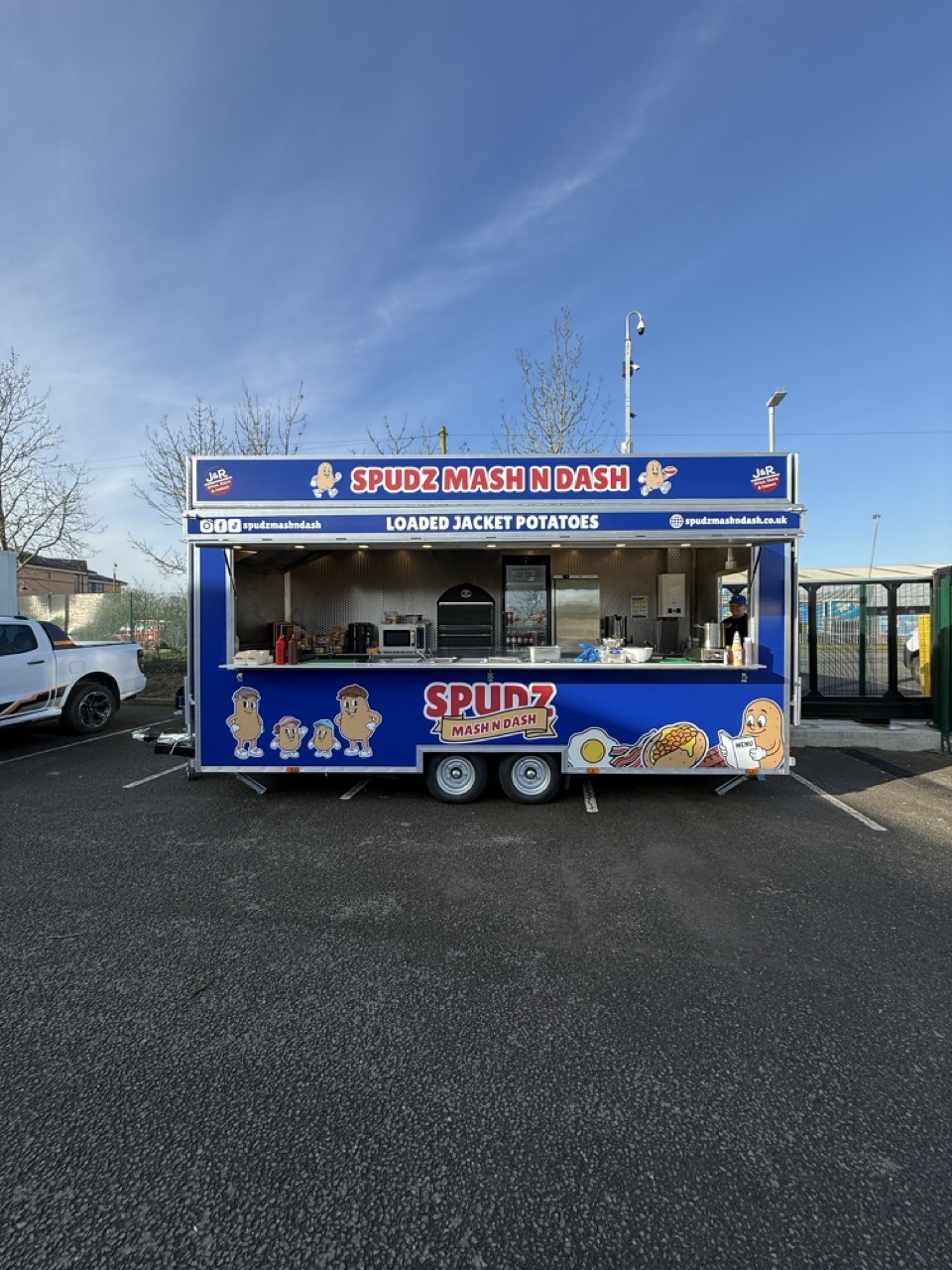 A blue food truck parked on a wet street with a smiling cartoon potato character holding a tray of food walking in front, with brick houses and a Union Jack flag in the background.