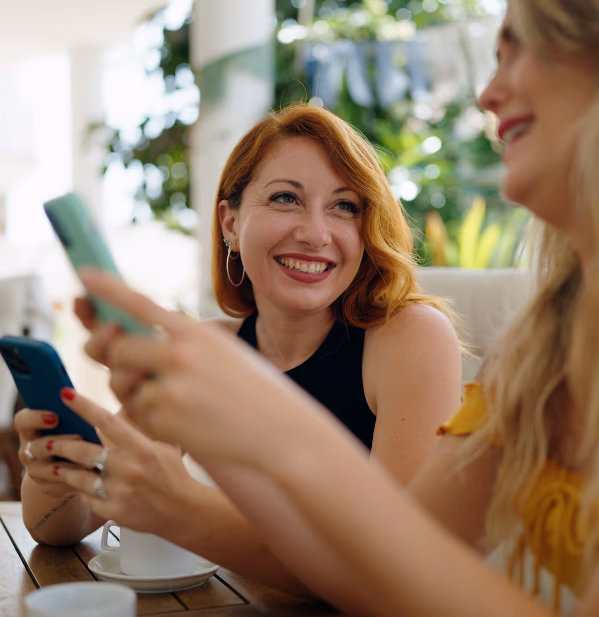 Deux femmes souriantes discutant dans un café, tenant chacune un smartphone.