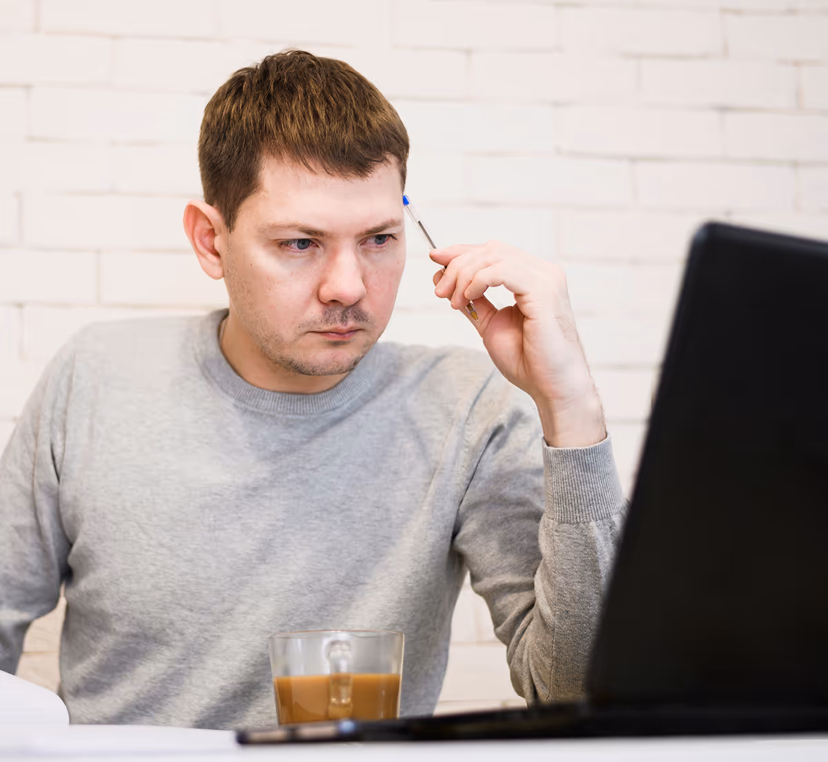 Homme pensif tenant un stylo bleu près de la tempe, regardant un ordinateur portable avec une tasse de café à côté.