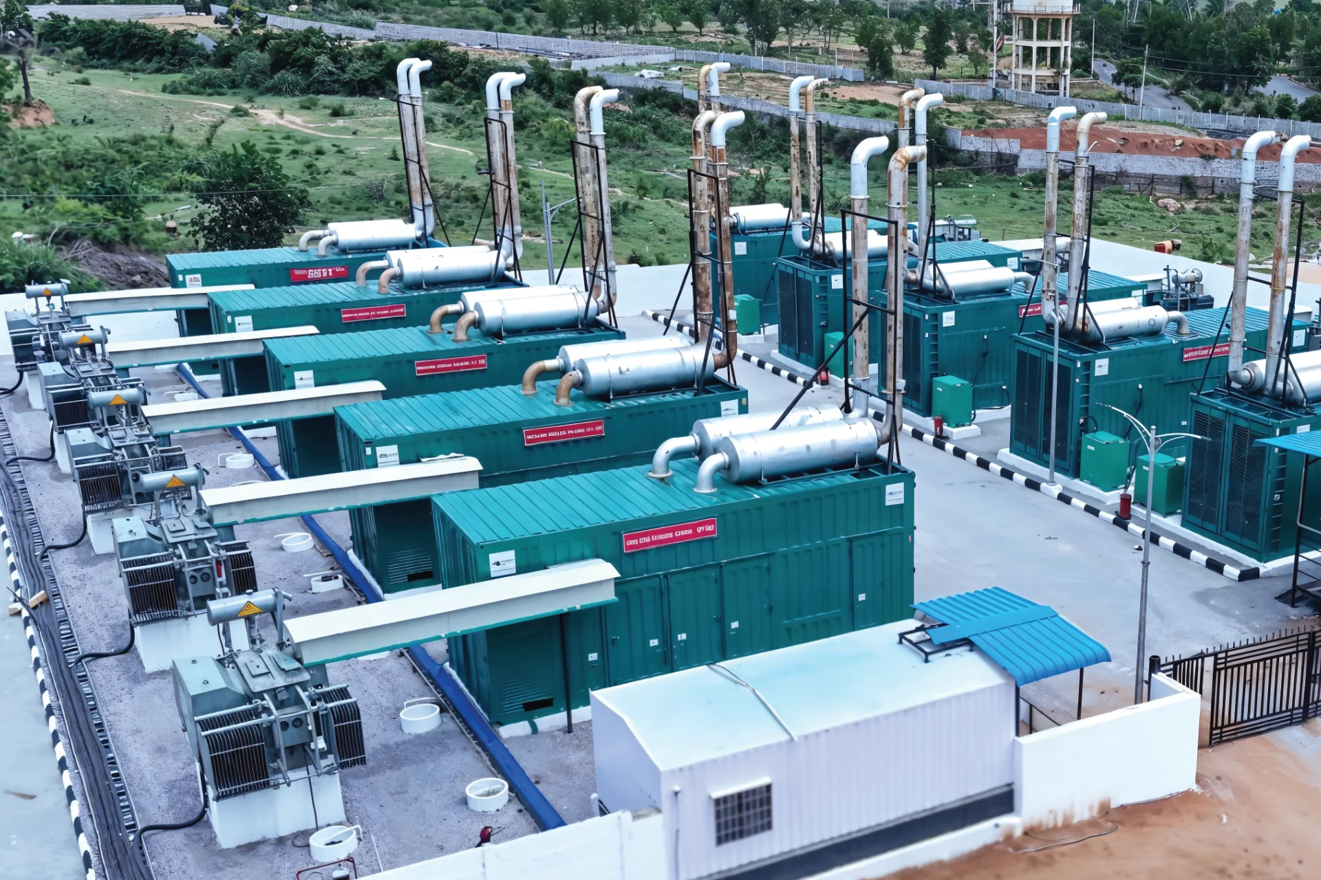 Industrial power generator units with large exhaust pipes arranged in rows at an outdoor facility with green equipment and surrounding greenery.