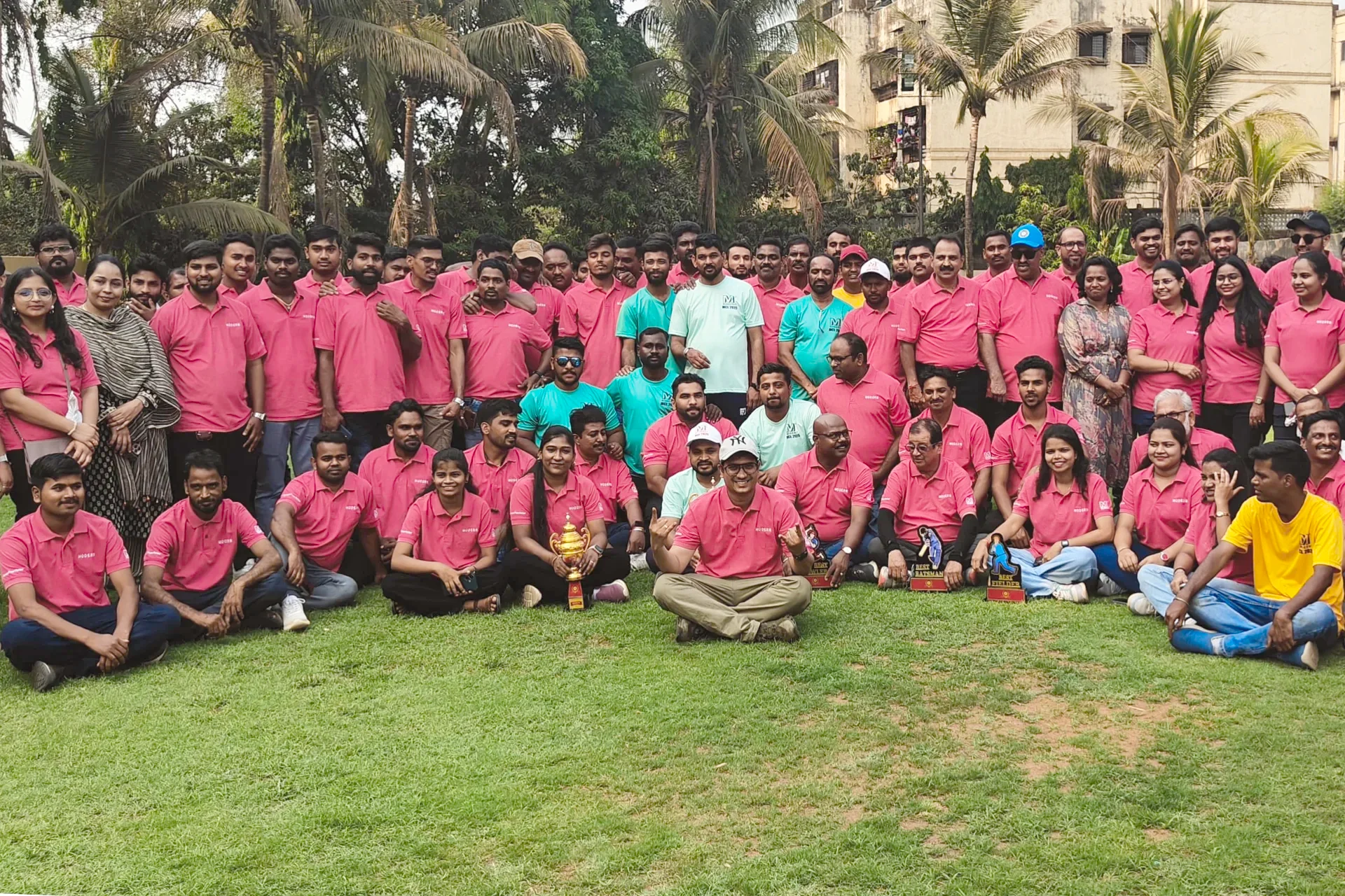 Large group of people outdoors on grass, mostly wearing pink polo shirts, some in teal and yellow shirts, with trophies displayed in front.