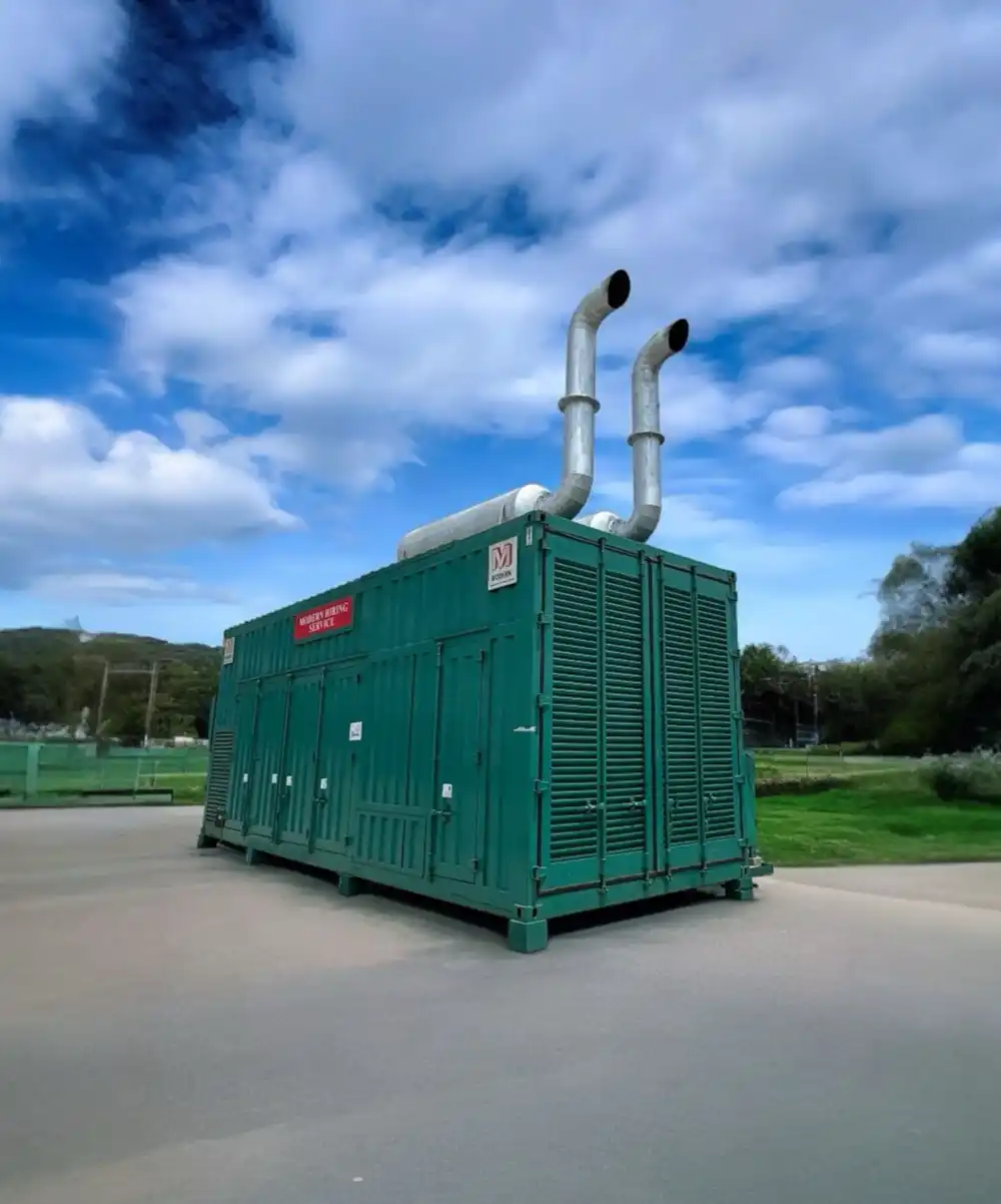 Large green industrial generator unit with two silver exhaust pipes on top, set on paved ground under a blue sky with scattered clouds.
