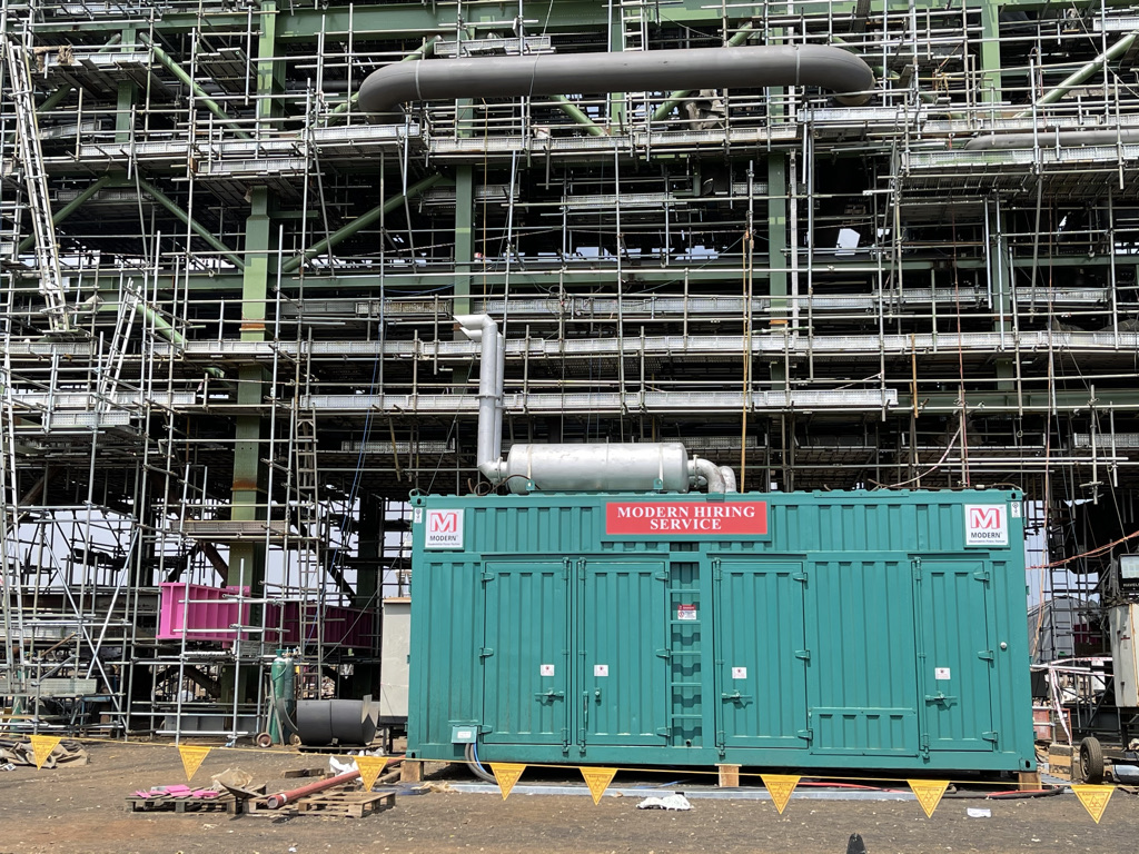 Green shipping container labeled 'Modern Hiring Service' in front of extensive scaffolding at a construction site.