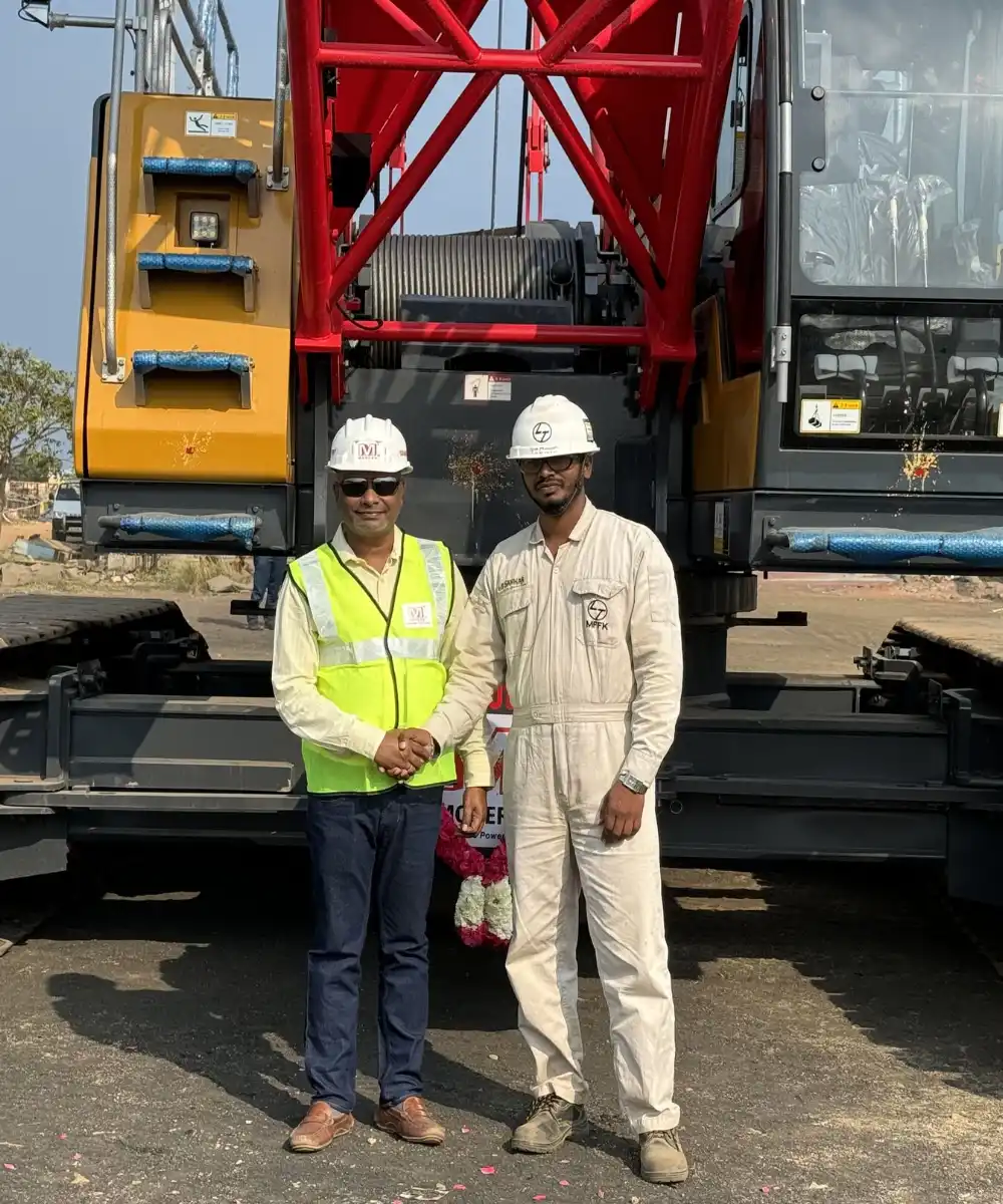 Two construction workers wearing hard hats shaking hands in front of a large red and yellow crane.