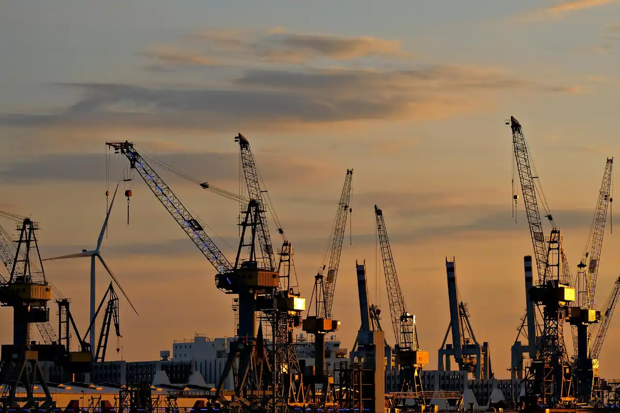 Silhouettes of numerous cranes and a wind turbine at a harbor during sunset with an orange sky.