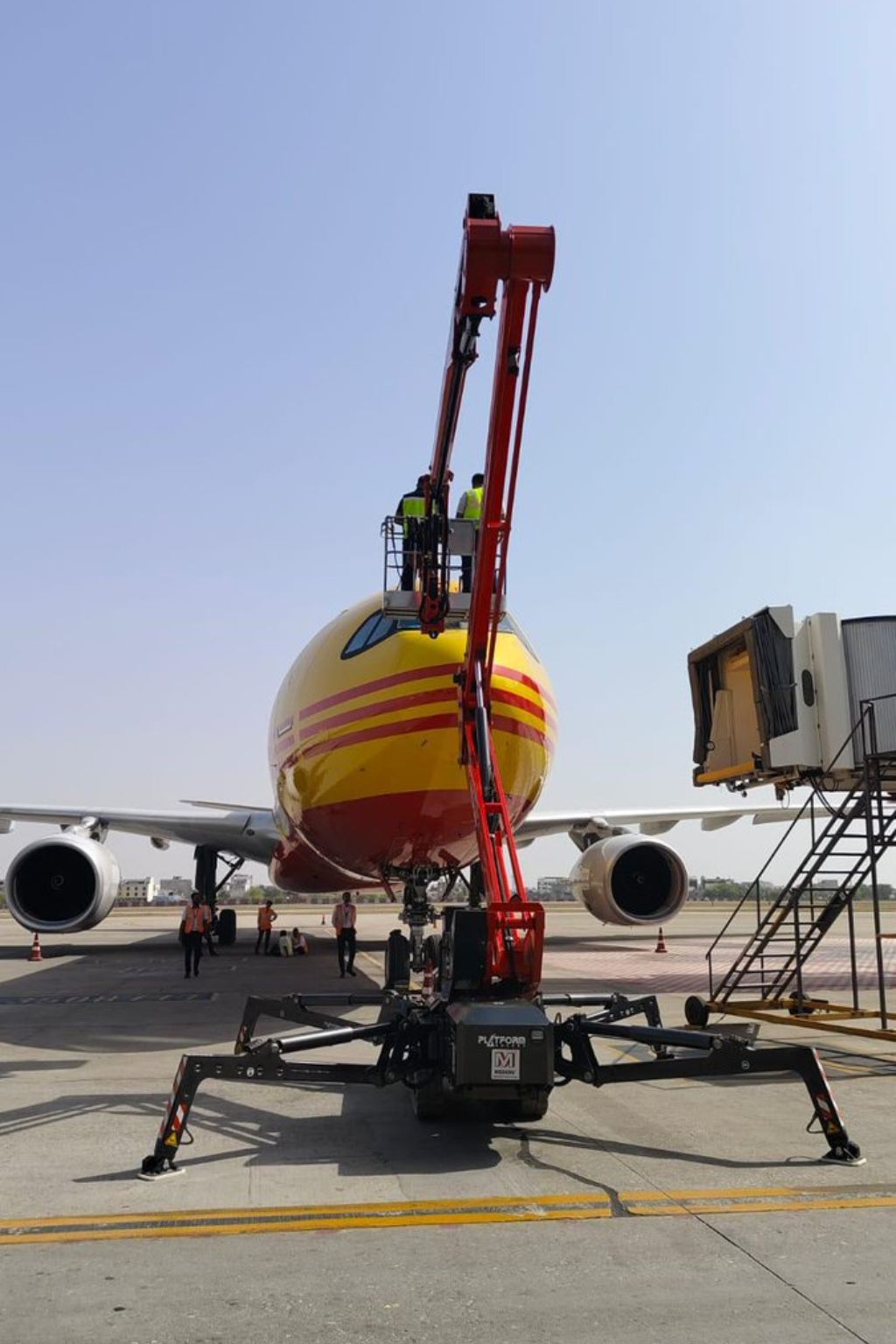 Front view of a yellow and red airplane on the tarmac with workers elevated on a red hydraulic platform inspecting the aircraft nose.