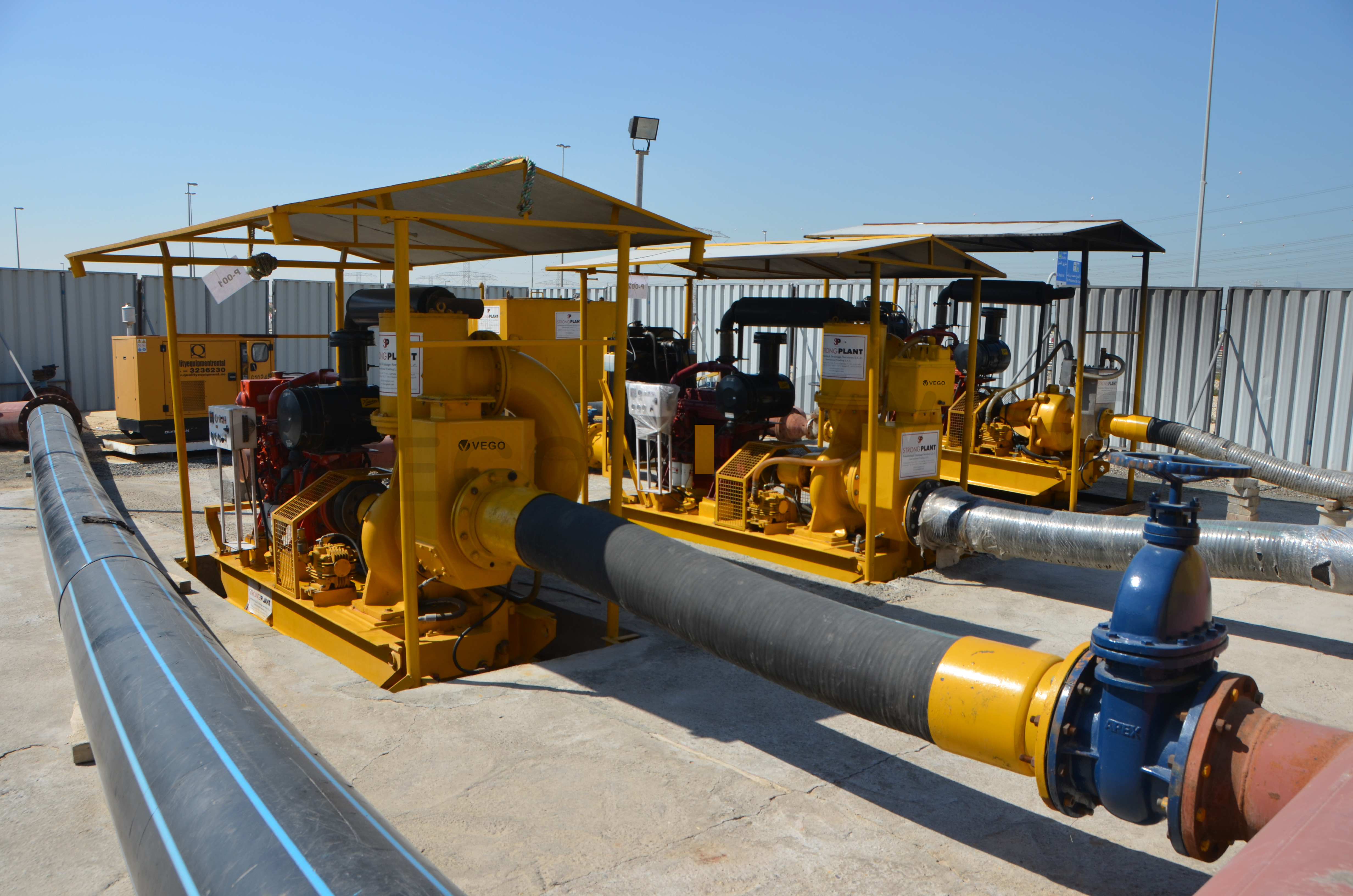 Three yellow industrial water pumps connected by large black hoses on a concrete surface under a clear blue sky.