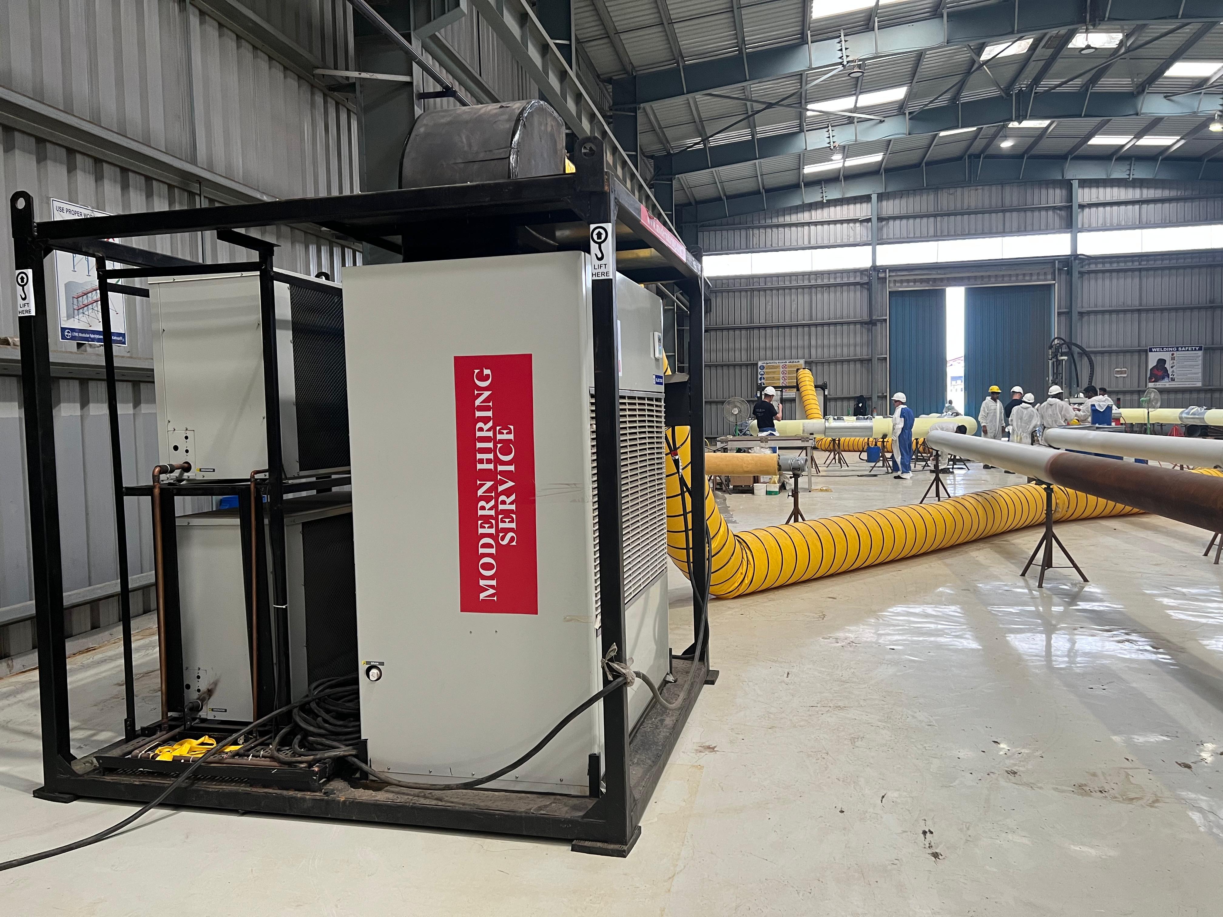 Industrial air conditioning unit with 'Modern Hiring Service' label inside a warehouse with workers and large ventilation ducts.