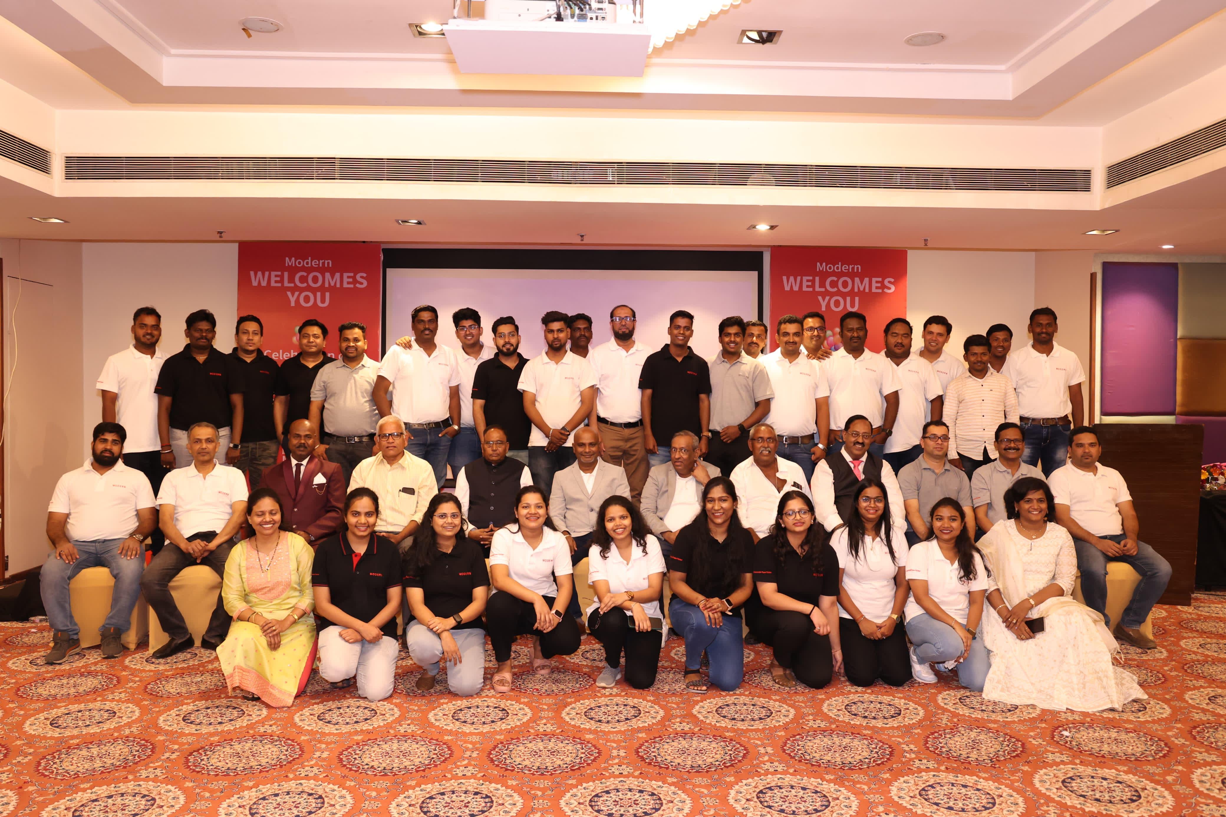 Smiling woman in glasses holding a tablet and making a fist pump gesture in a modern office setting with colleagues in the background.
