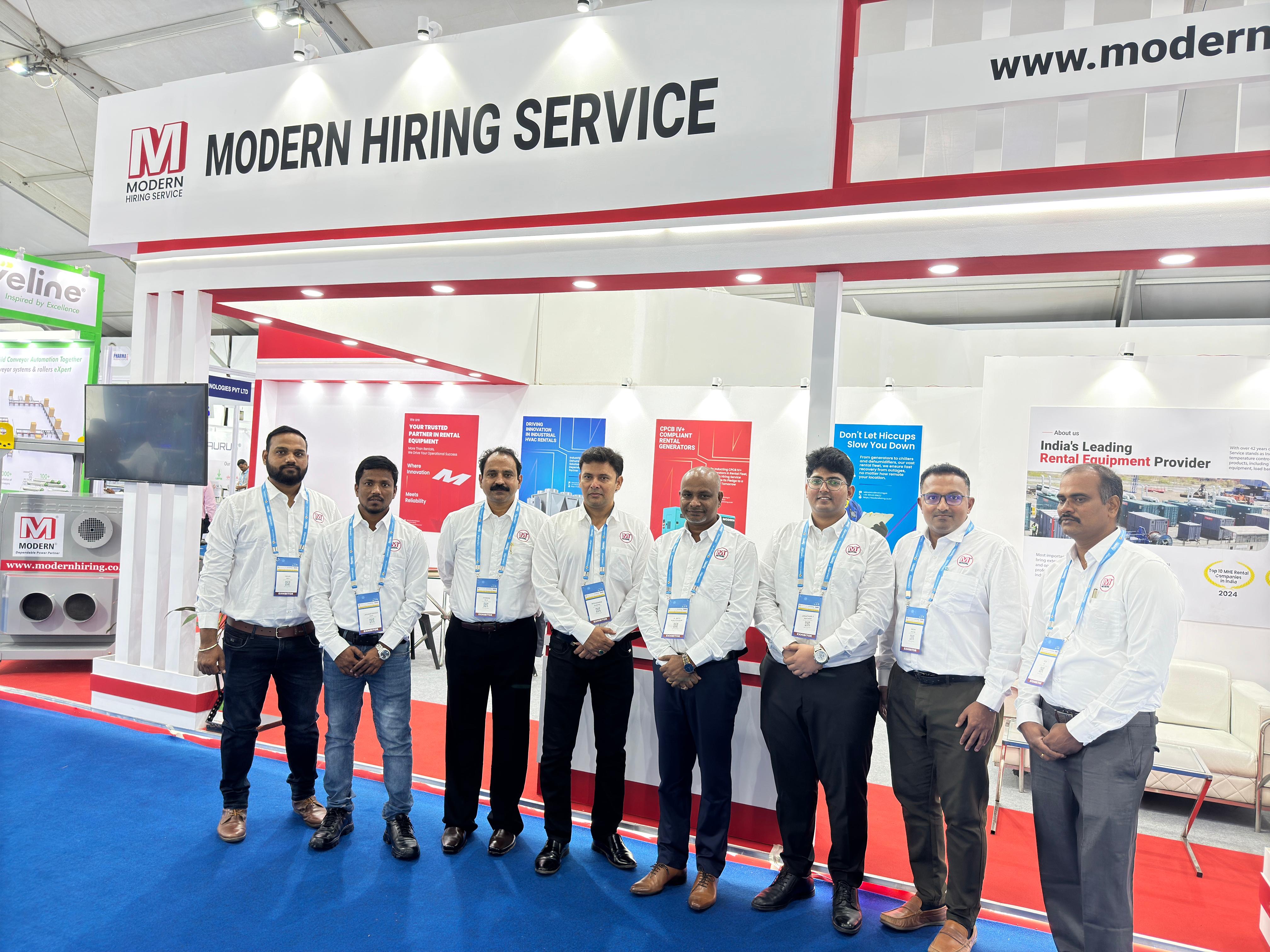 Seven men in business attire posing at a Modern Hiring Service booth with red and white signage inside an exhibition hall.