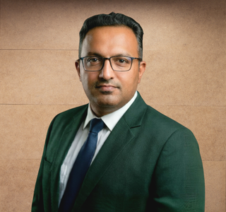 Man with glasses wearing a green suit, white shirt, and blue tie against a brown background.