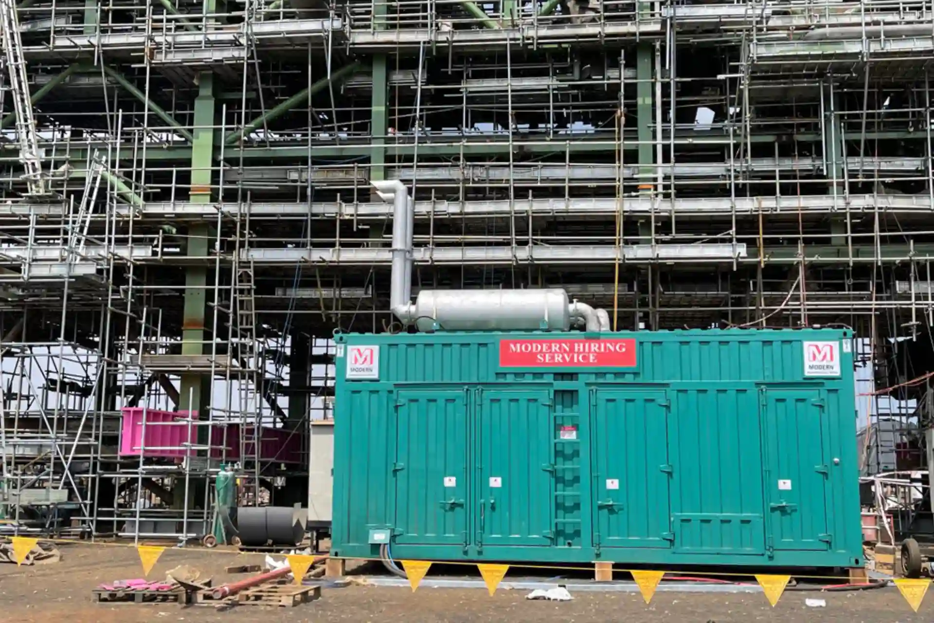 Green shipping container labeled 'Modern Hiring Service' in front of extensive scaffolding at a construction site.