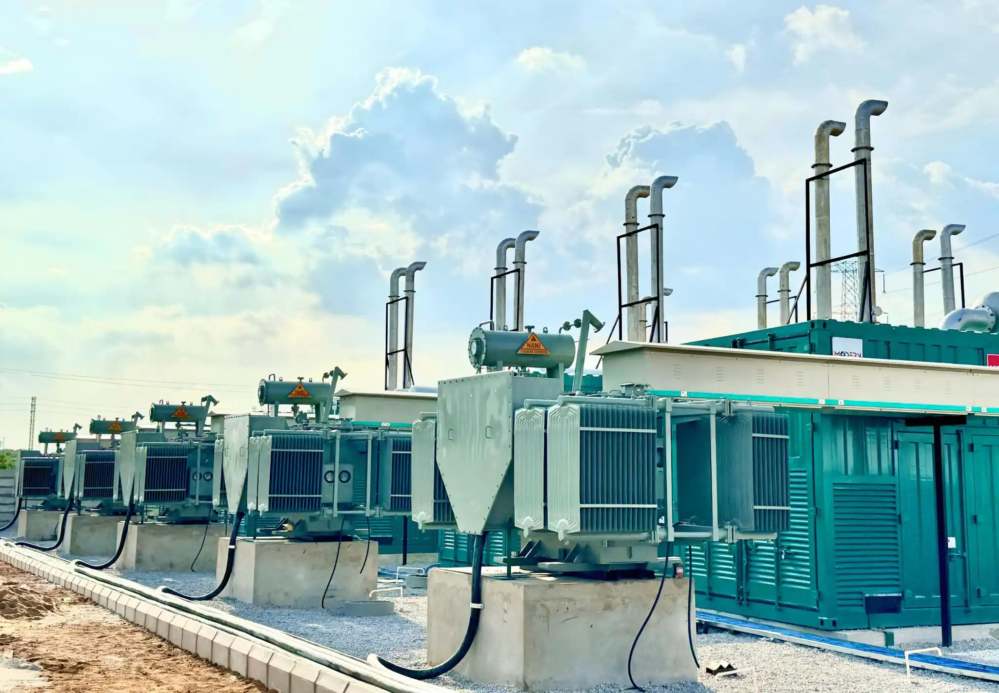Row of industrial electrical transformers with tall exhaust pipes under a cloudy sky.