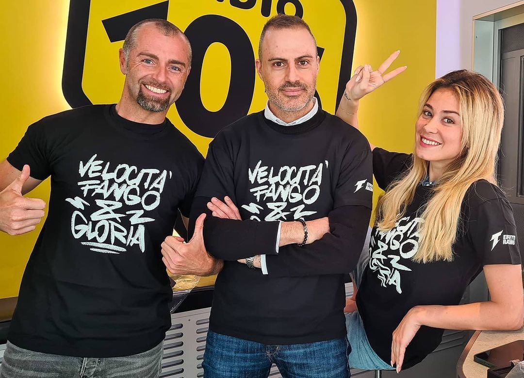 Three smiling people wearing black shirts with white text posing in front of a yellow wall with black text.