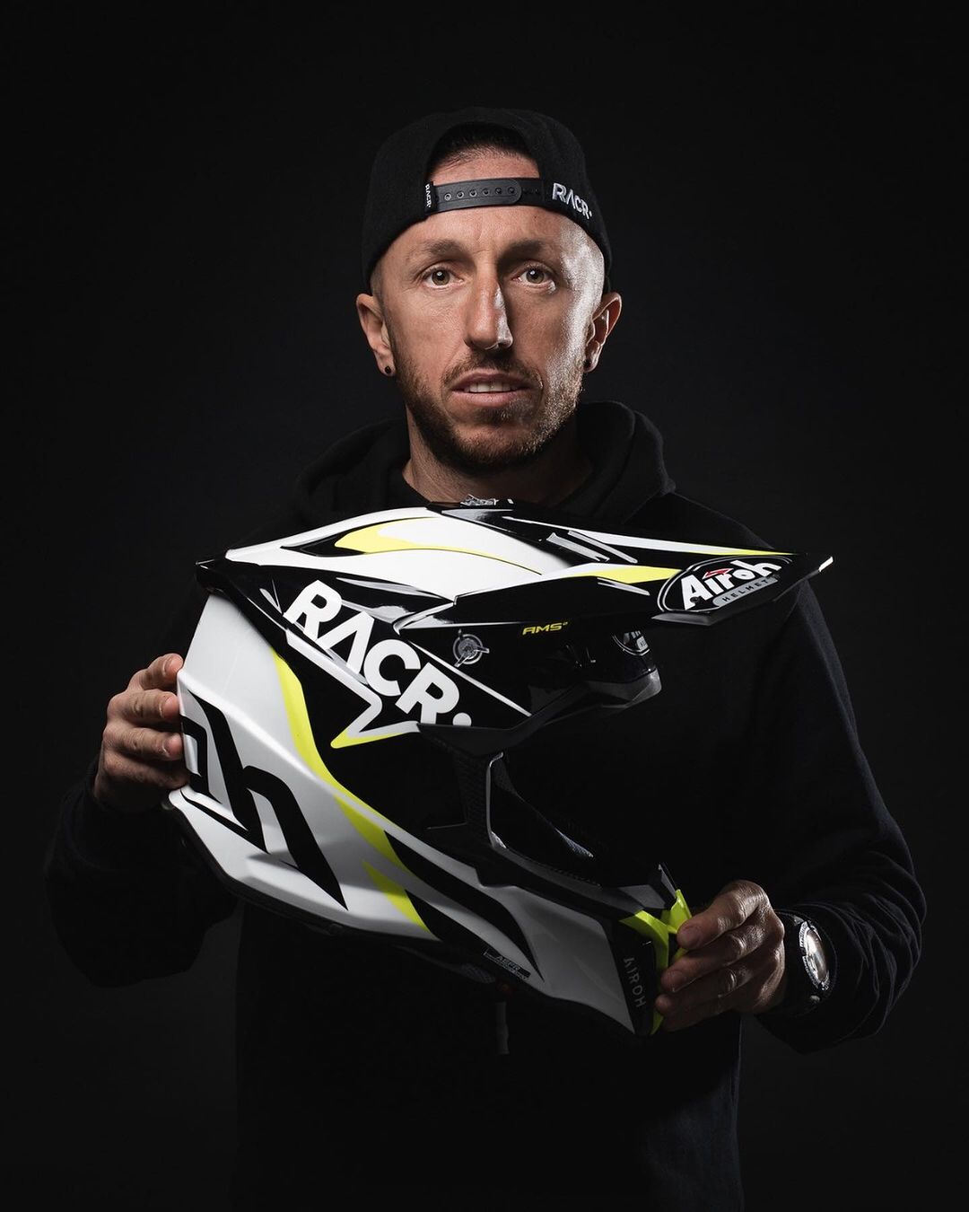 Man wearing a black hoodie and backwards cap holding a white, black, and yellow Airoh motocross helmet.