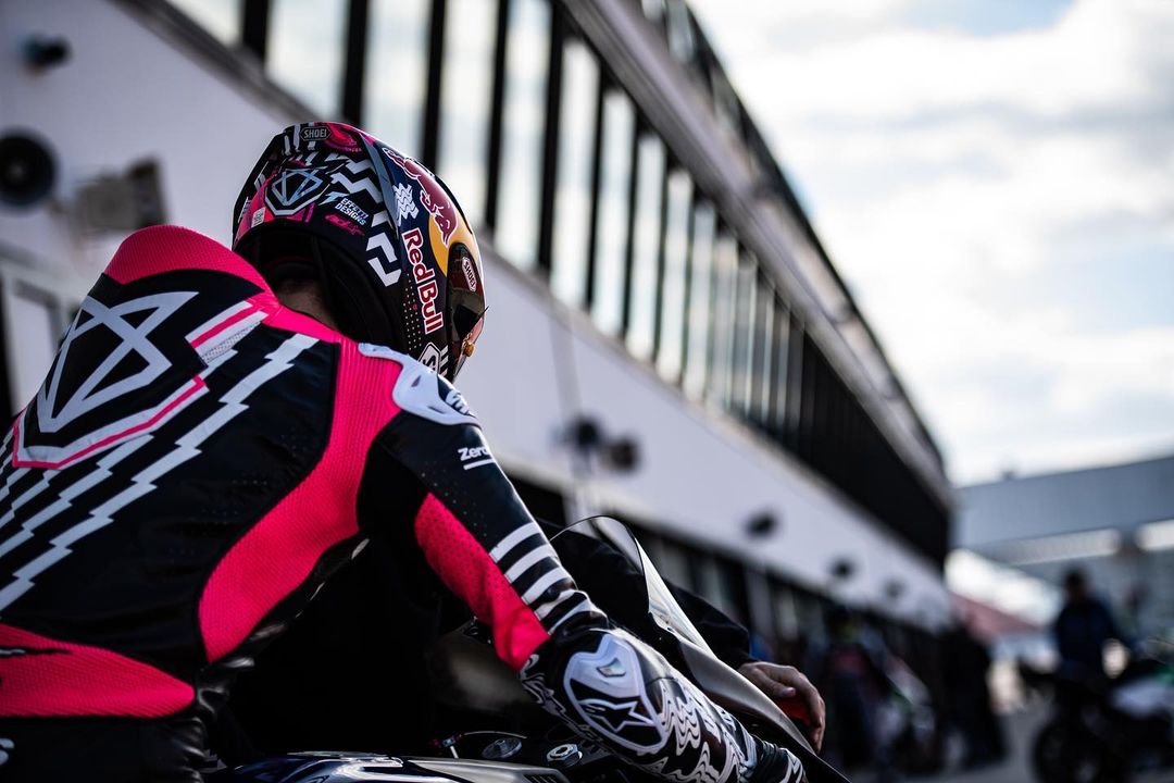 Motorcyclist in pink and black racing gear with a Red Bull helmet sitting on a motorcycle near a modern race track building.