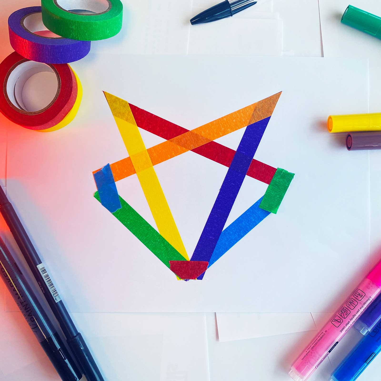 A white paper with a multicolored star made from overlapping strips of colored tape, surrounded by rolls of colored tape and markers.
