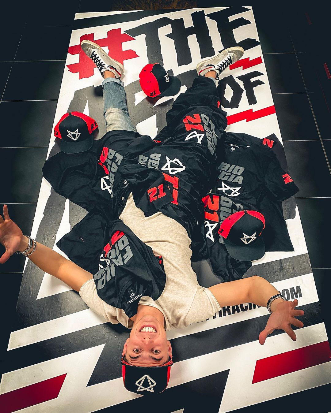 Person lying on a large banner wearing a black and red cap, surrounded by black merchandise shirts and caps with 'Diggia' branding.