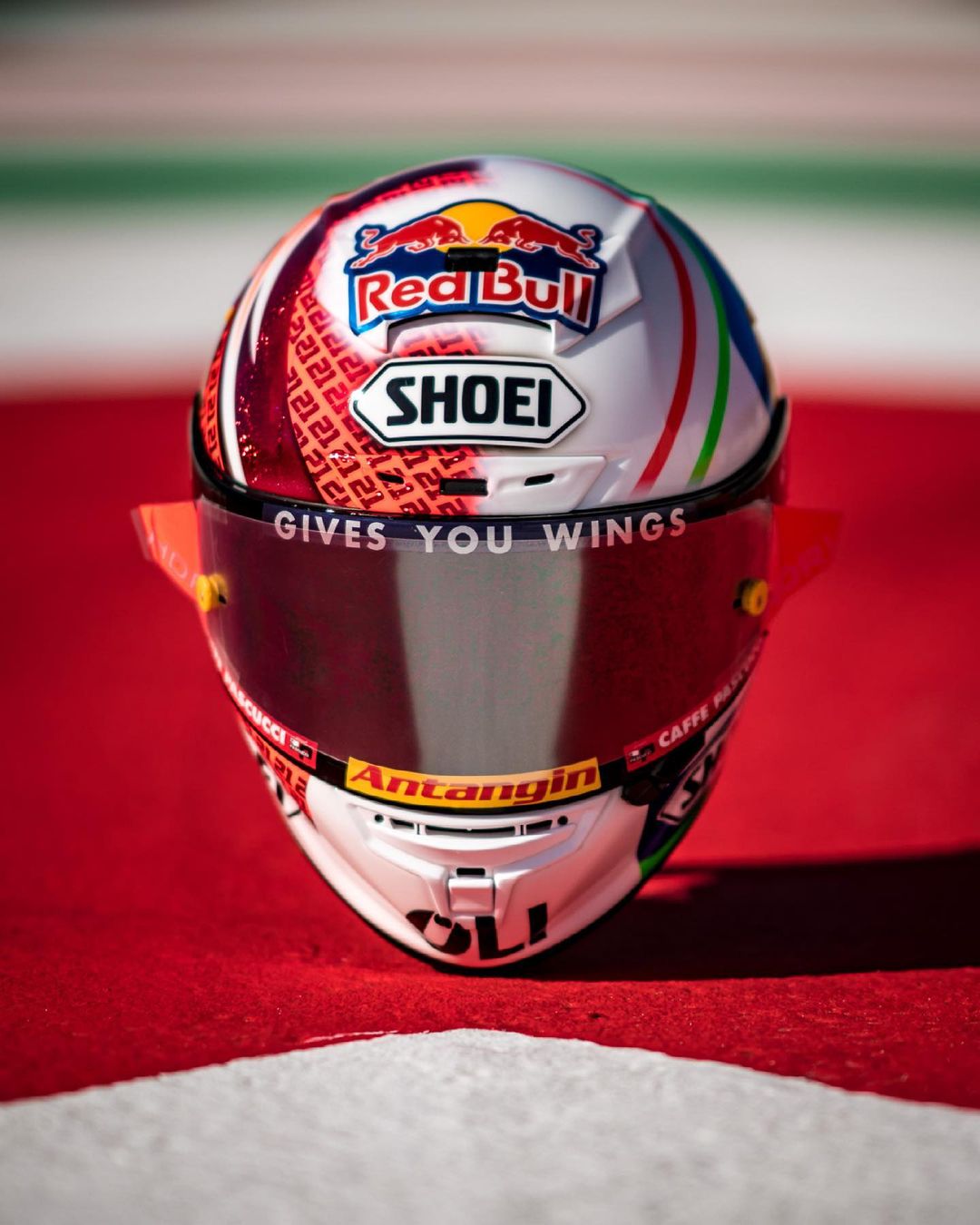Front view of a white racing helmet with Red Bull, Shoei, and Antangin logos, placed on red and white racetrack surface.