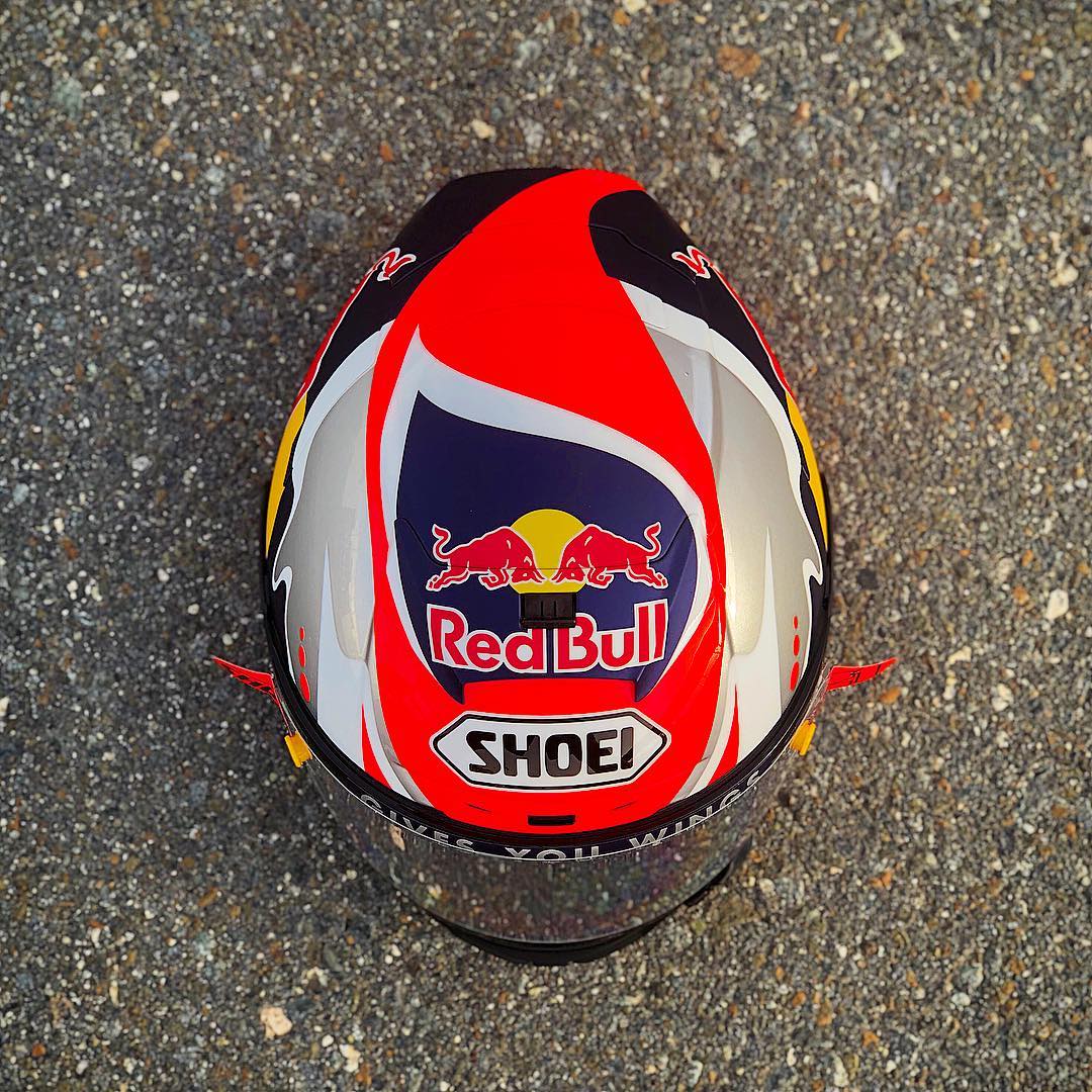 Top view of a red, white, yellow, and black motorcycle helmet with Red Bull and Shoei logos on a rough asphalt surface.