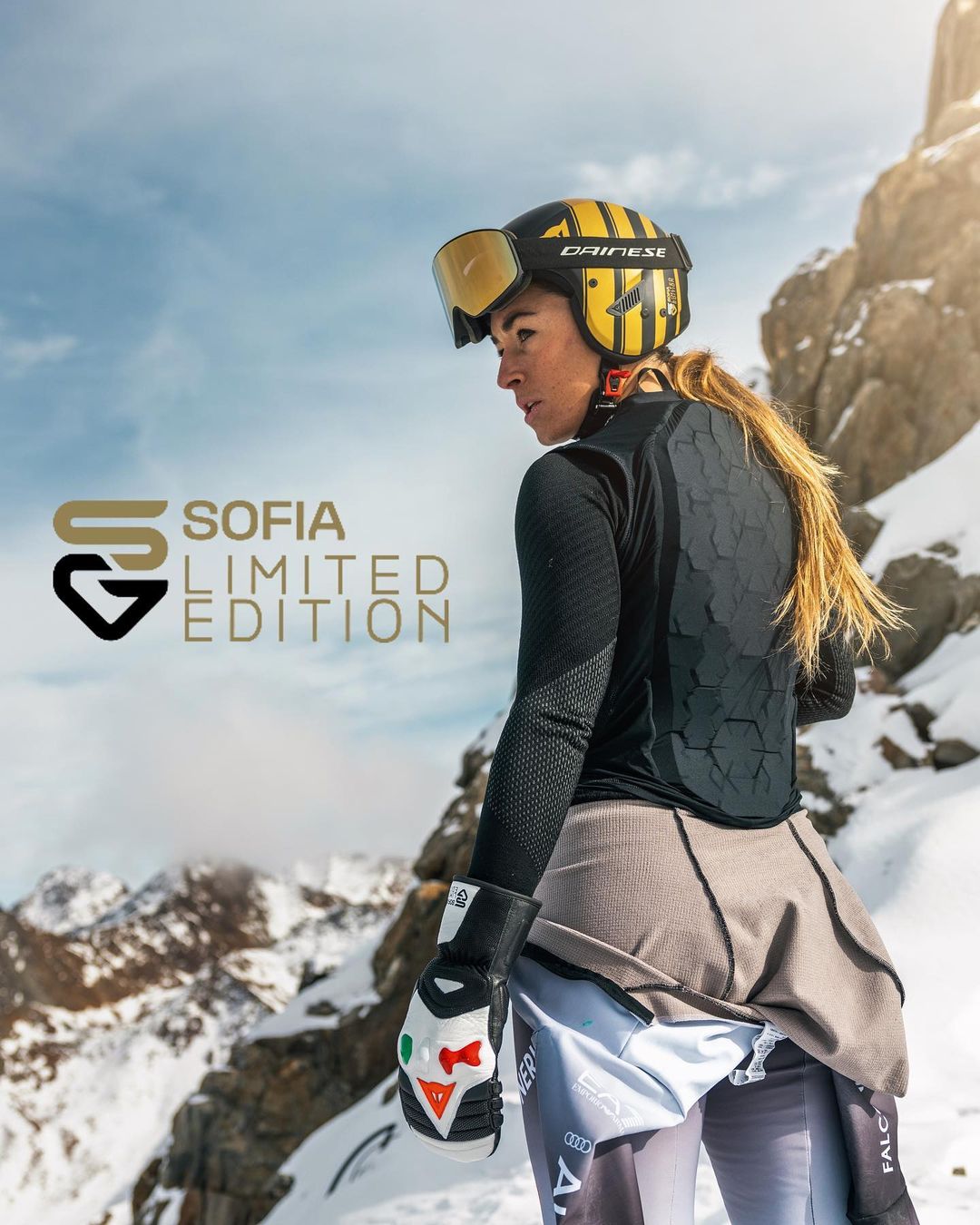 Female skier with long braided hair wearing a yellow and black helmet and black gloves, standing in snowy mountain terrain with rocky peaks, beside text reading 'Sofia Limited Edition'.