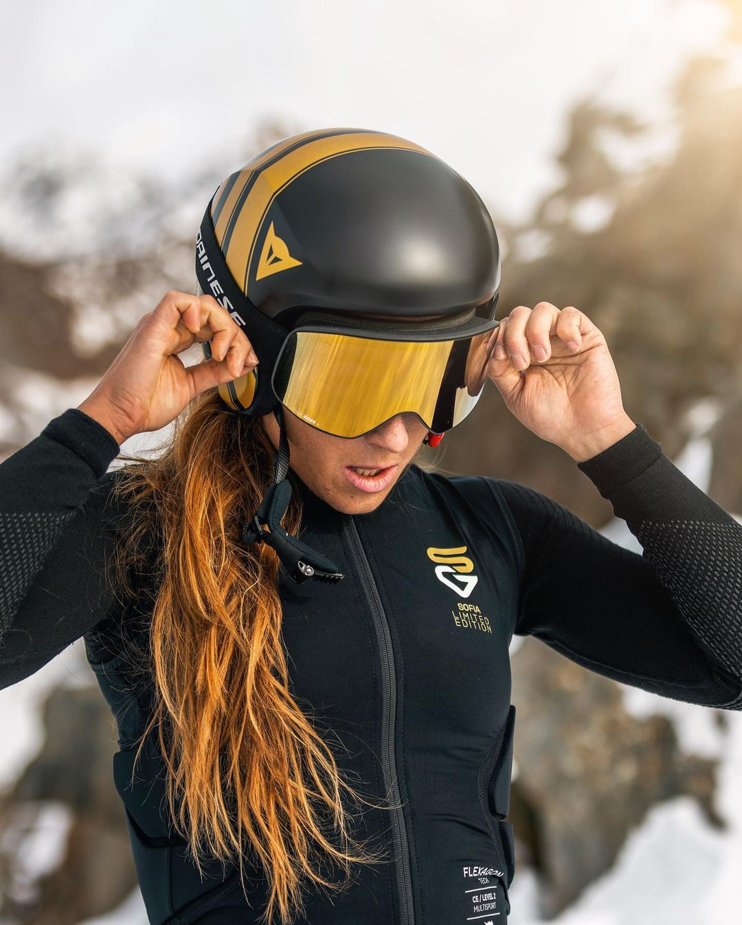 Person with long red hair adjusting a black and gold helmet with reflective yellow goggles, wearing a black sports jacket labeled Sofia Limited Edition.
