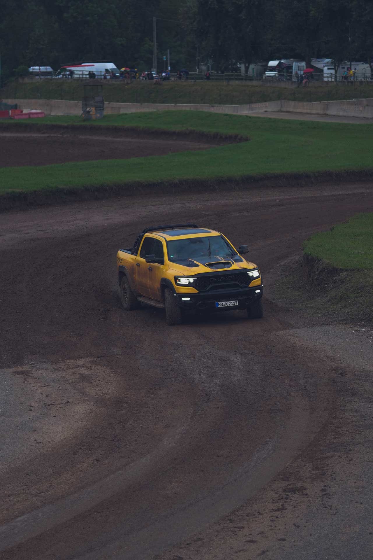 Yellow RAM pickup truck driving on a muddy dirt race track with grass borders and spectators in the background.