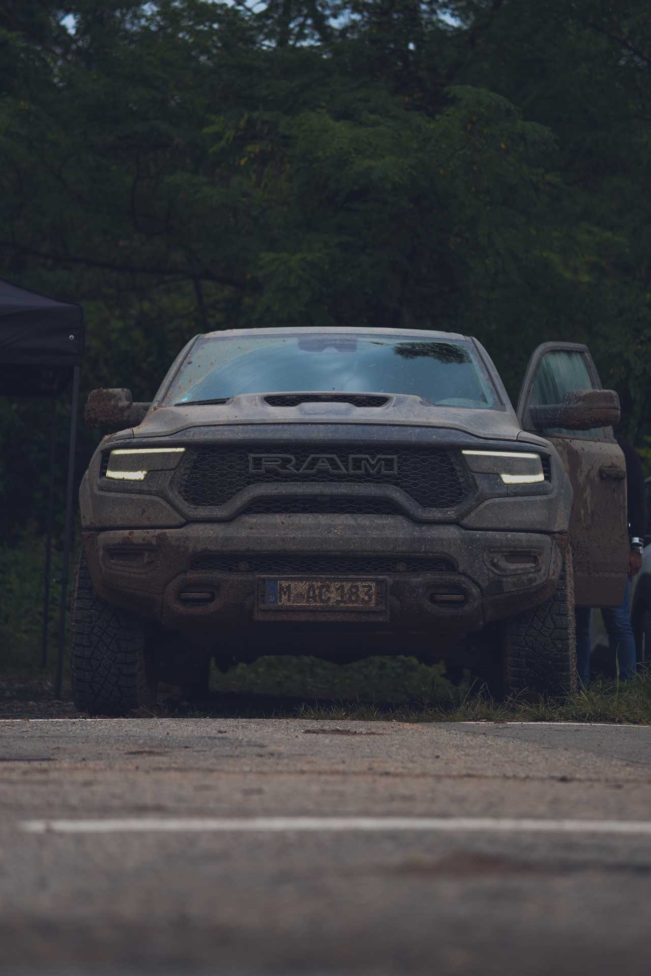 Front view of a muddy RAM truck with headlights on and the driver's door open, parked on a path with trees in the background.