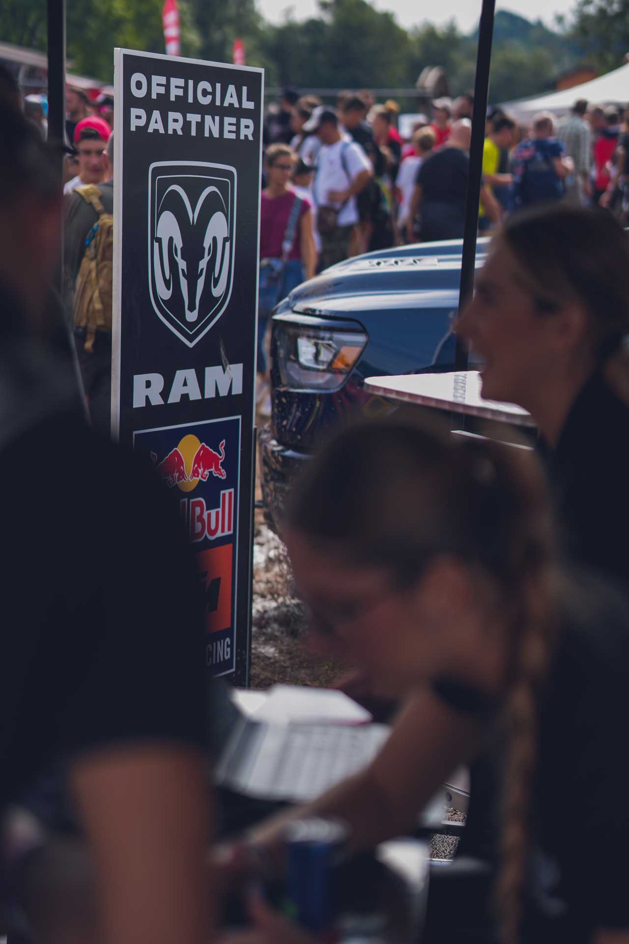 Signboard showing RAM as an official partner with Red Bull and KTM logos at an outdoor event with people and vehicles in the background.