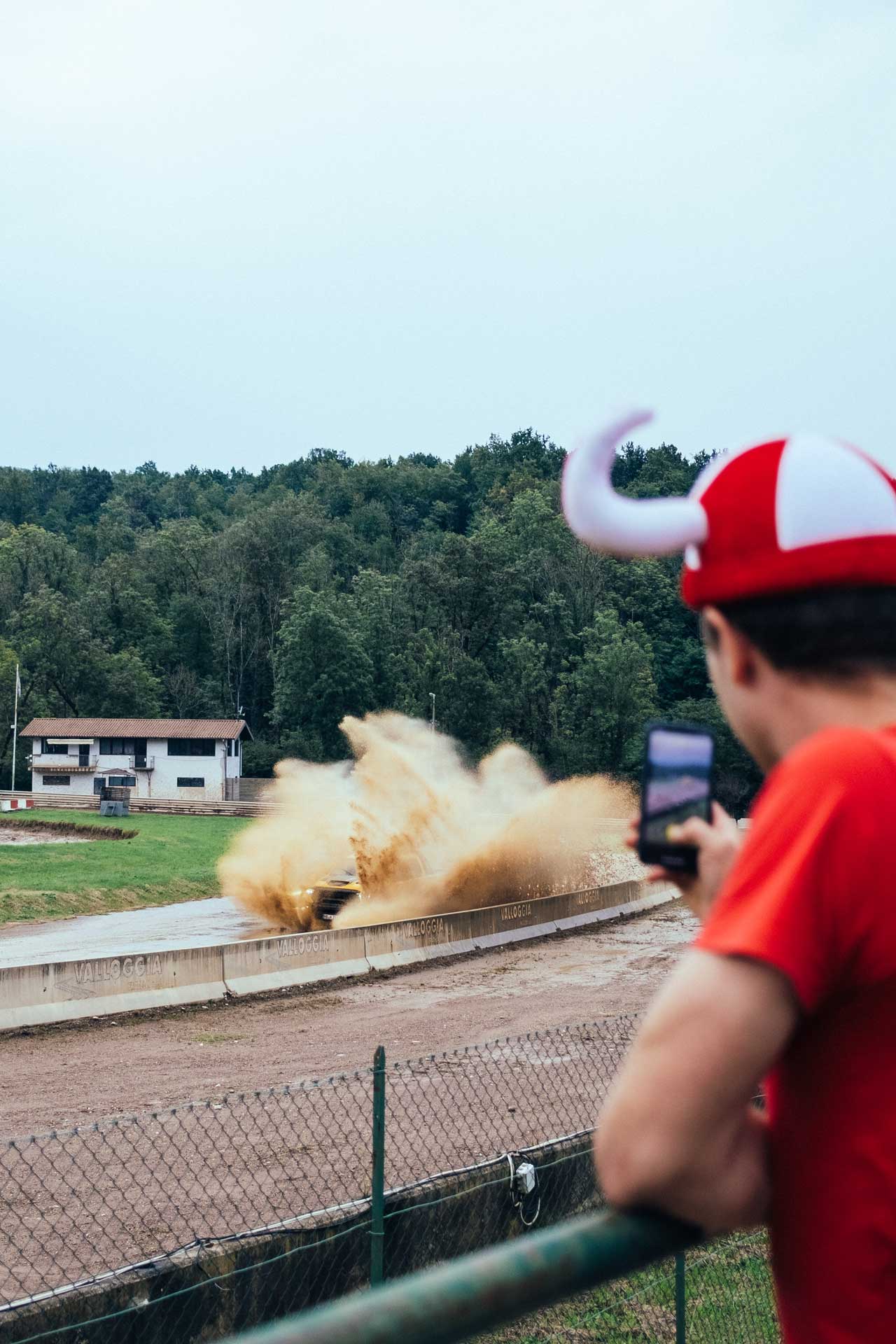 Person wearing red shirt and horned hat filming a car kicking up dirt on a race track.
