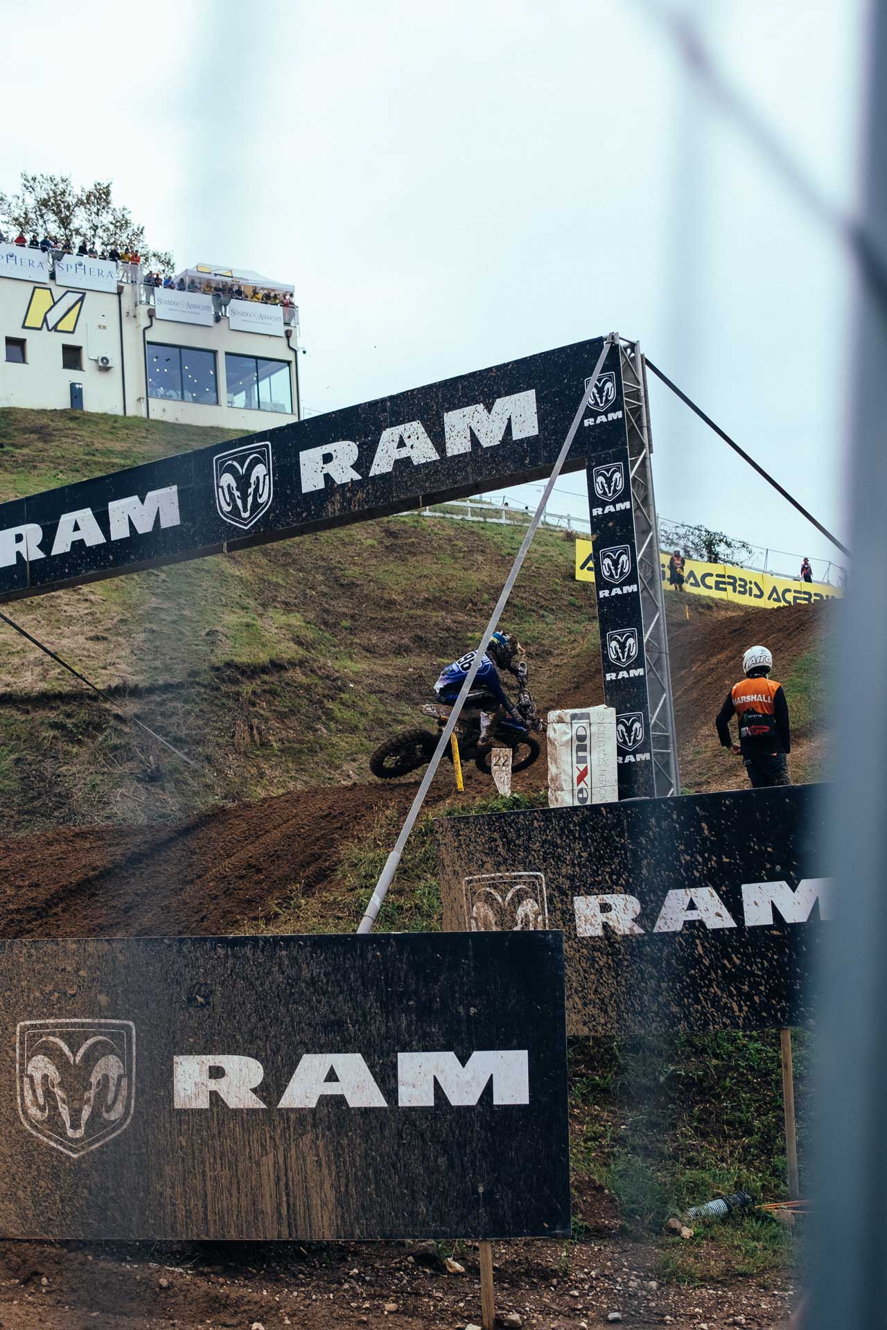 Motocross rider in blue gear navigating a muddy dirt track under large RAM sponsor banners.