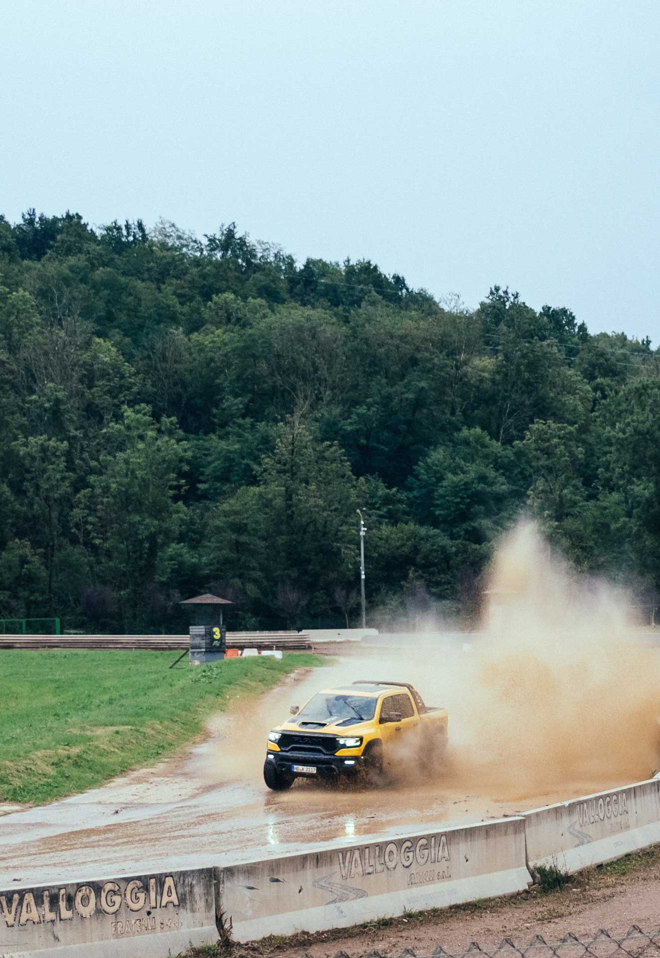 Yellow pickup truck driving fast on a wet dirt track, kicking up a large spray of mud.