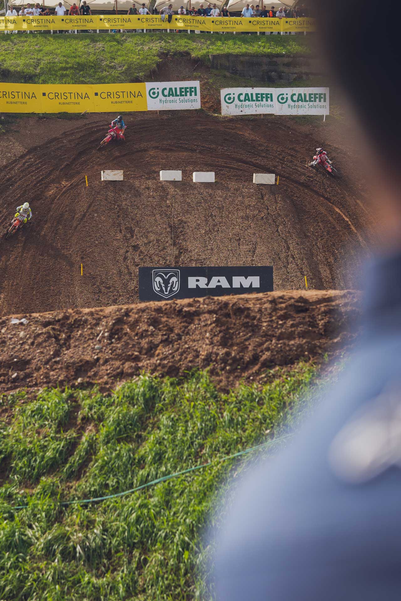 Three motocross riders racing on a dirt track with banners for RAM, Cristina, and Caleffi in the background and spectators behind a barrier.
