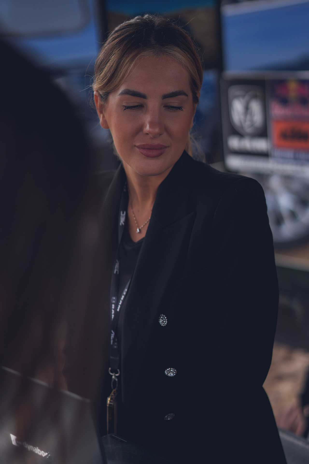 Portrait of a woman with closed eyes wearing a black blazer and a lanyard, in a softly lit indoor setting.