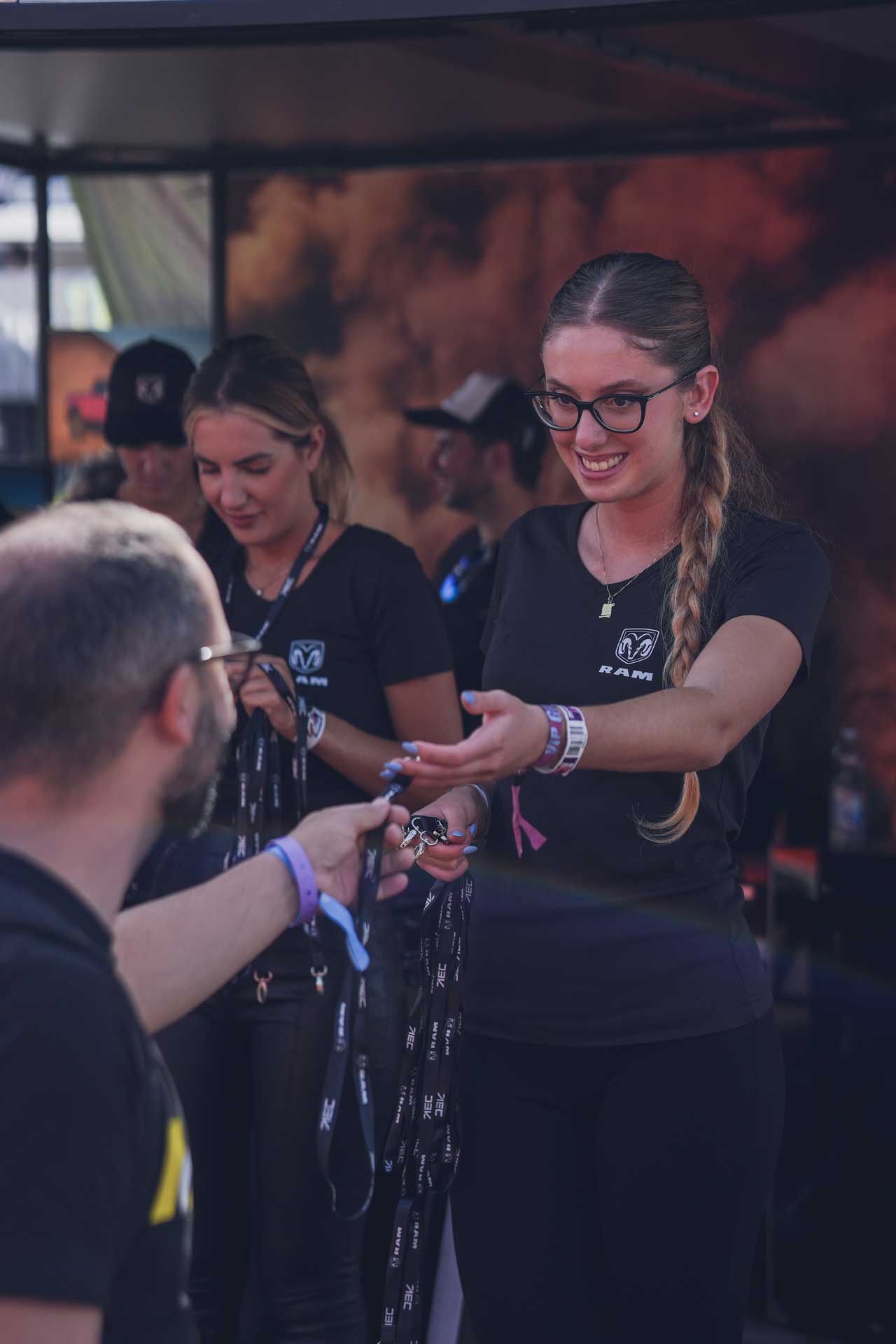 A smiling woman with glasses and a braid hands a lanyard to a man while another woman in a RAM shirt holds more lanyards in the background.