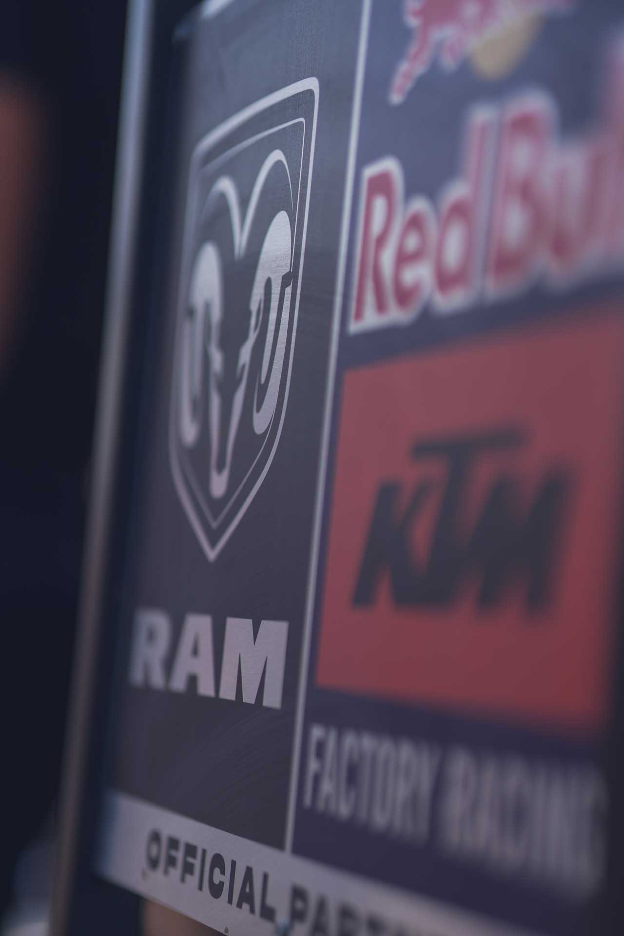 Close-up of logos including RAM, Red Bull, and KTM Factory Racing on a racing vehicle or equipment.