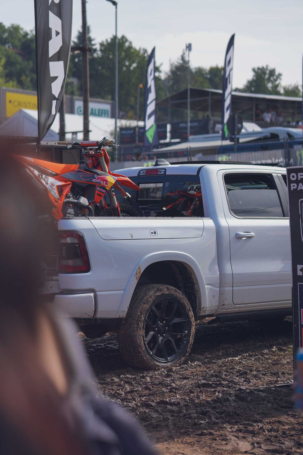White pickup truck with mud-covered tires carrying an orange motocross bike on a muddy track at an outdoor event.