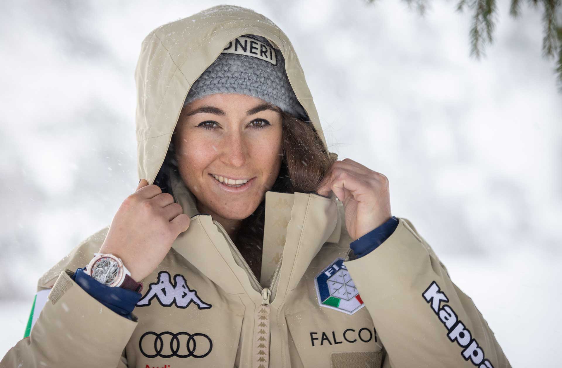 Smiling woman outdoors in snow wearing a beige jacket with ski-related patches, a gray knit hat, and holding the jacket hood.