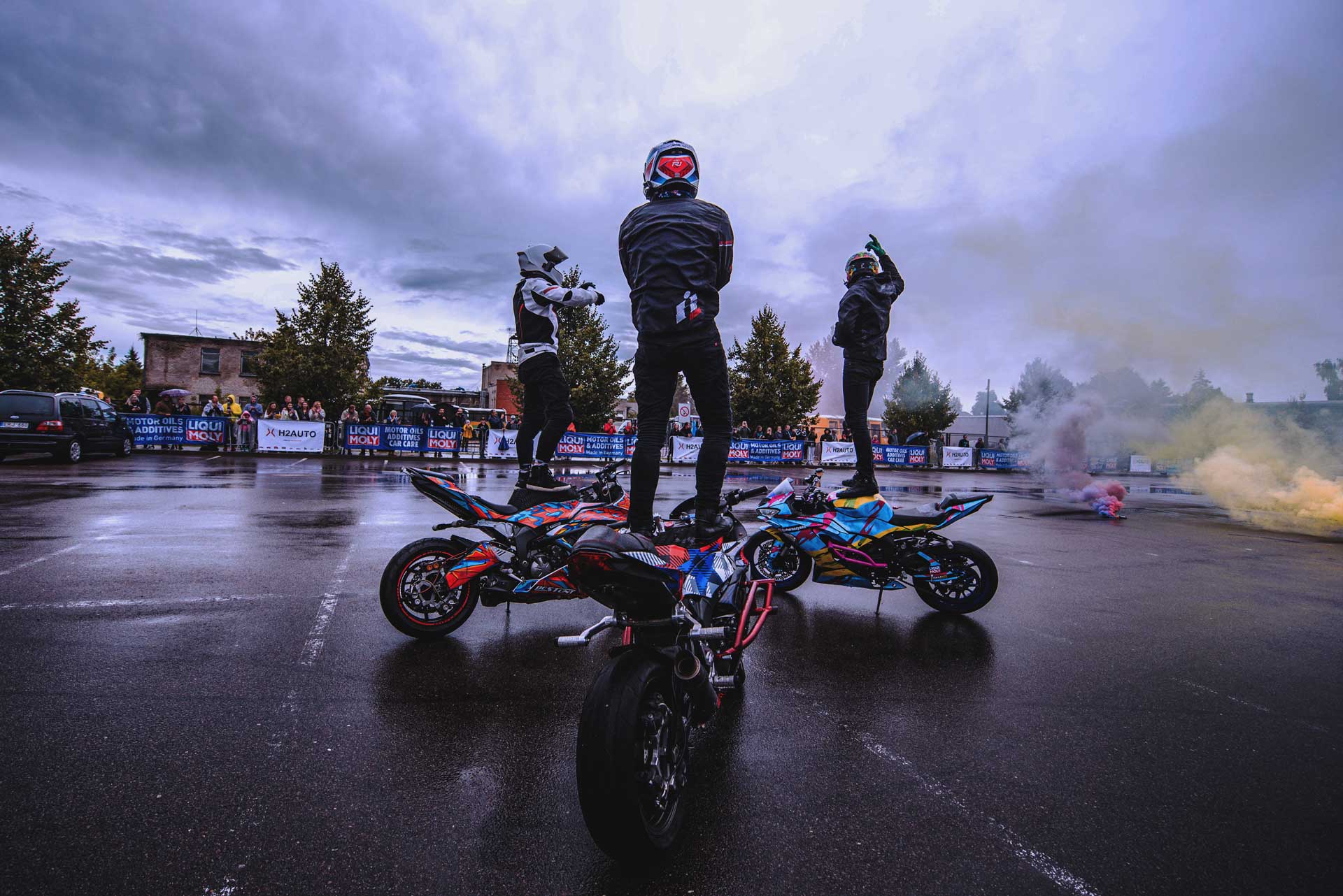 Three motorcyclists in helmets and protective gear standing on their colorful sport motorcycles in a wet parking lot with spectators and colored smoke in the background.