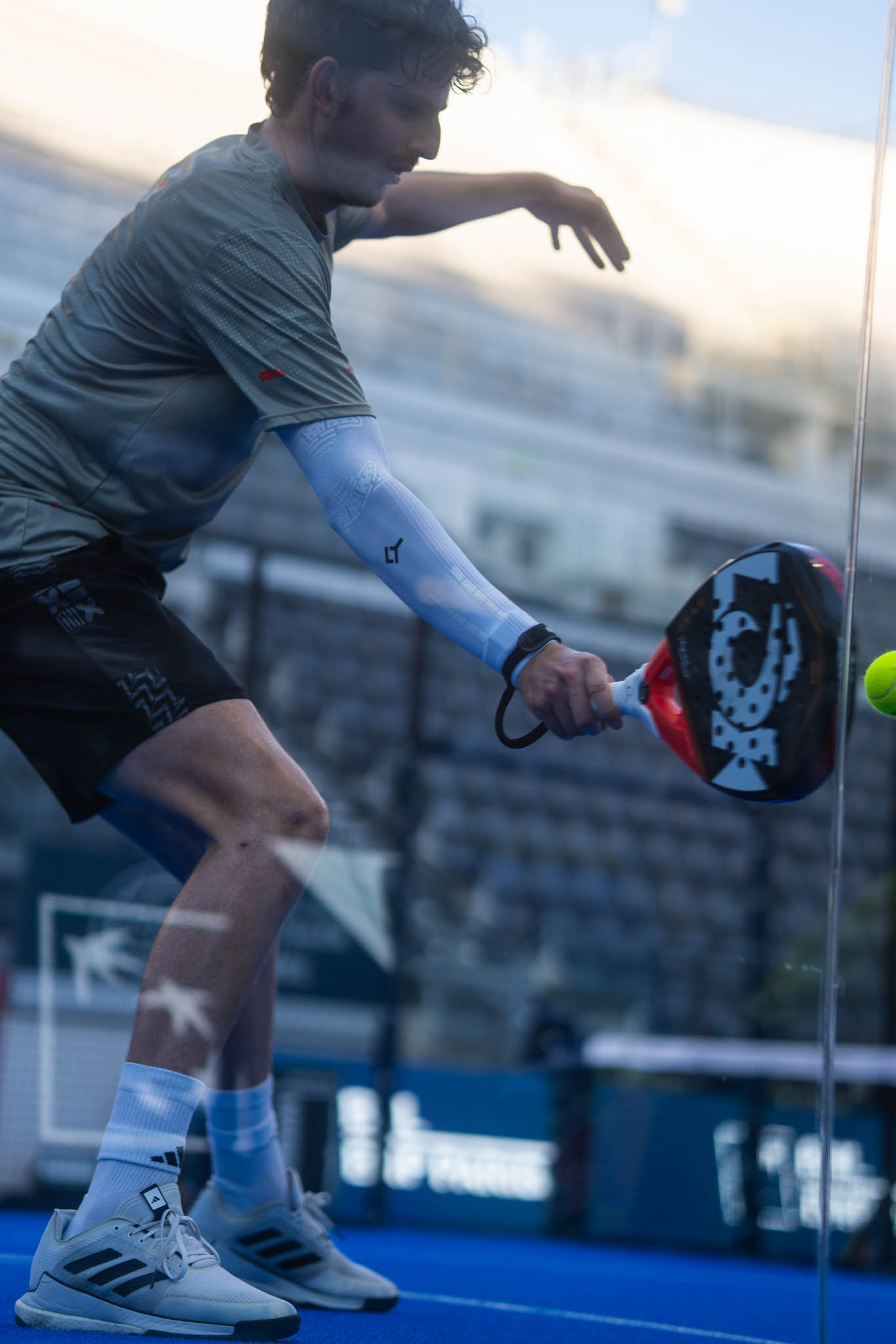 Man in sportswear playing padel, hitting a ball with a paddle on a blue court with a glass wall.