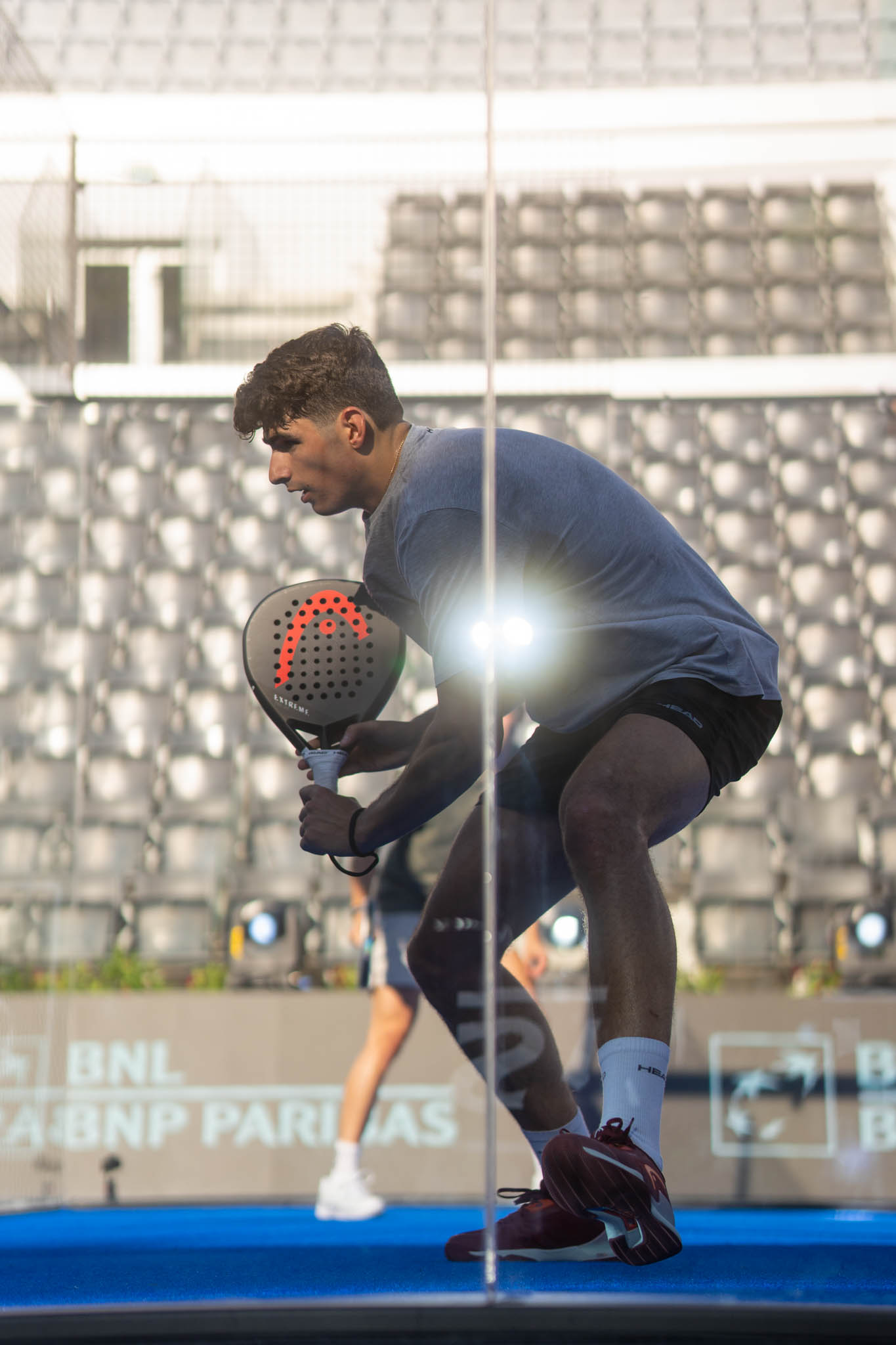 Man in a blue shirt and black shorts playing padel on a blue court, holding a racket and crouching in a ready position.
