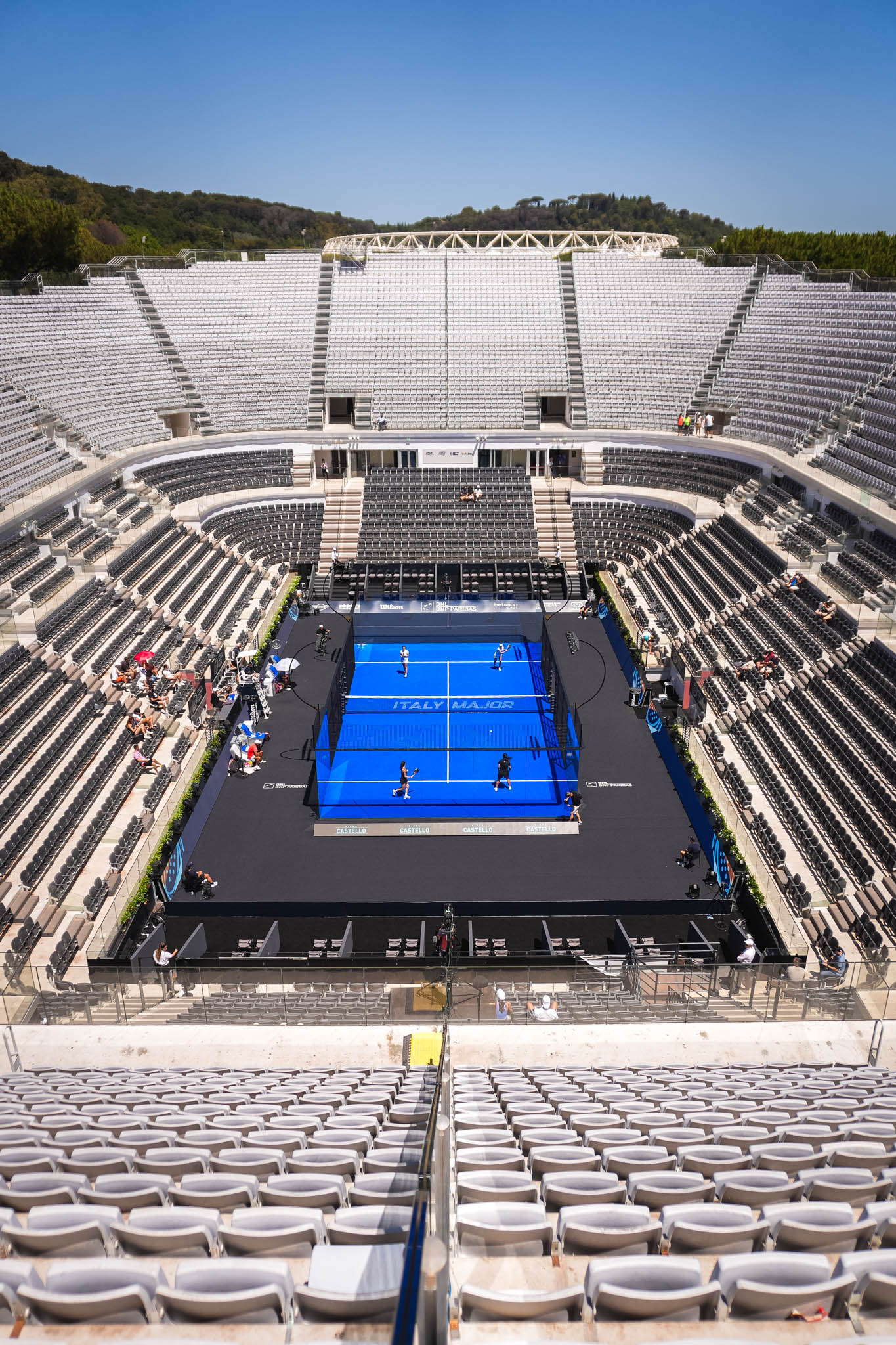 Empty large stadium with a blue padel court in the center and few people sitting and walking around.