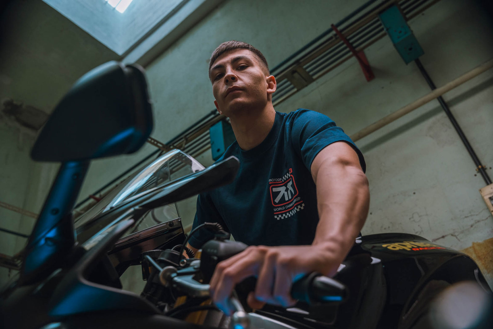 Man in a dark t-shirt sitting on a black motorcycle indoors, looking confidently at the camera.